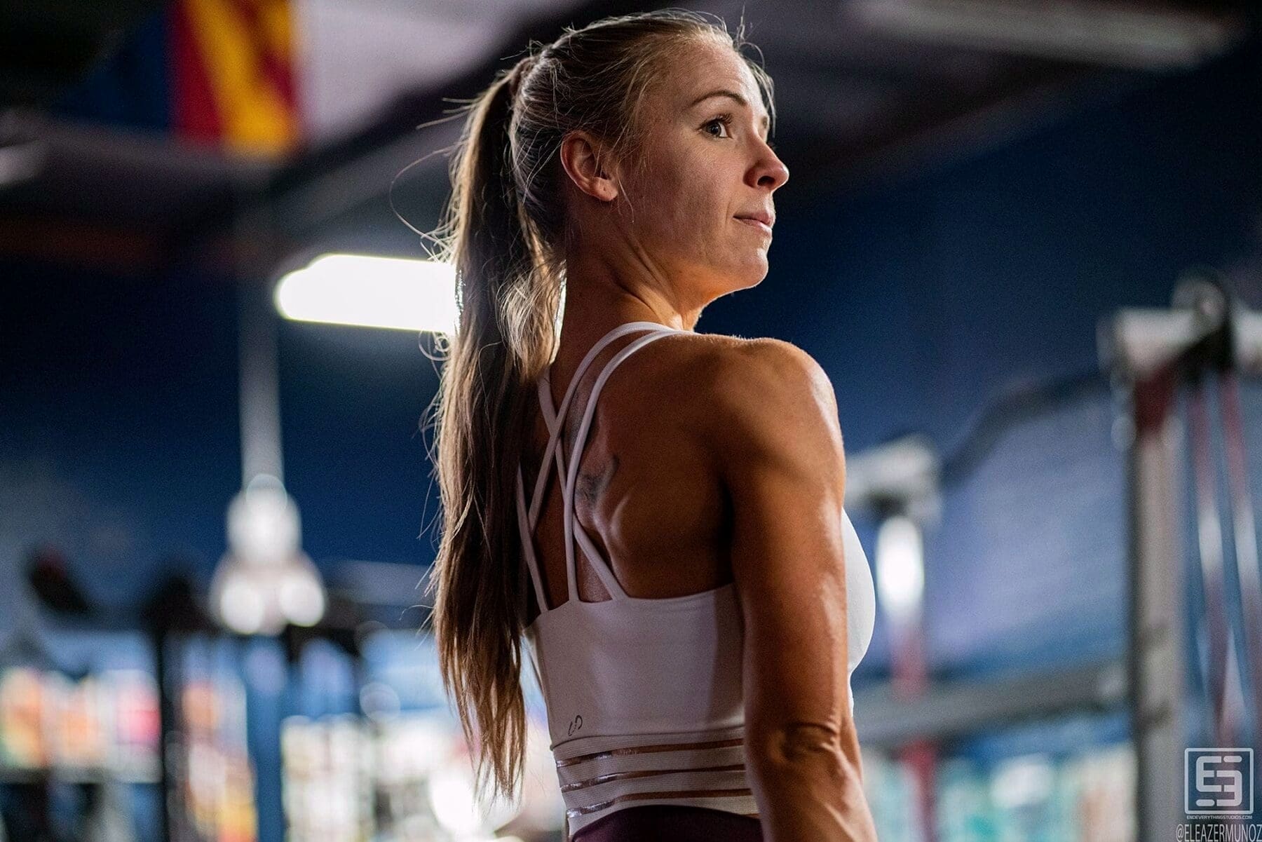 A fitness model looks back over her shoulder beneath directional gym lighting.