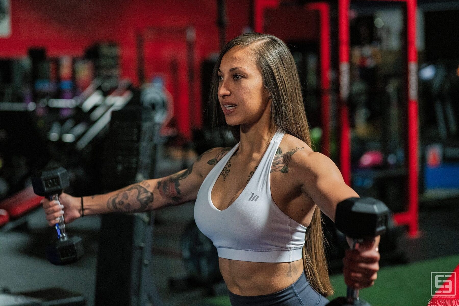 Danni lifts dumbbells to the sides during a shoulder exercise in a three-quarter gym portrait.