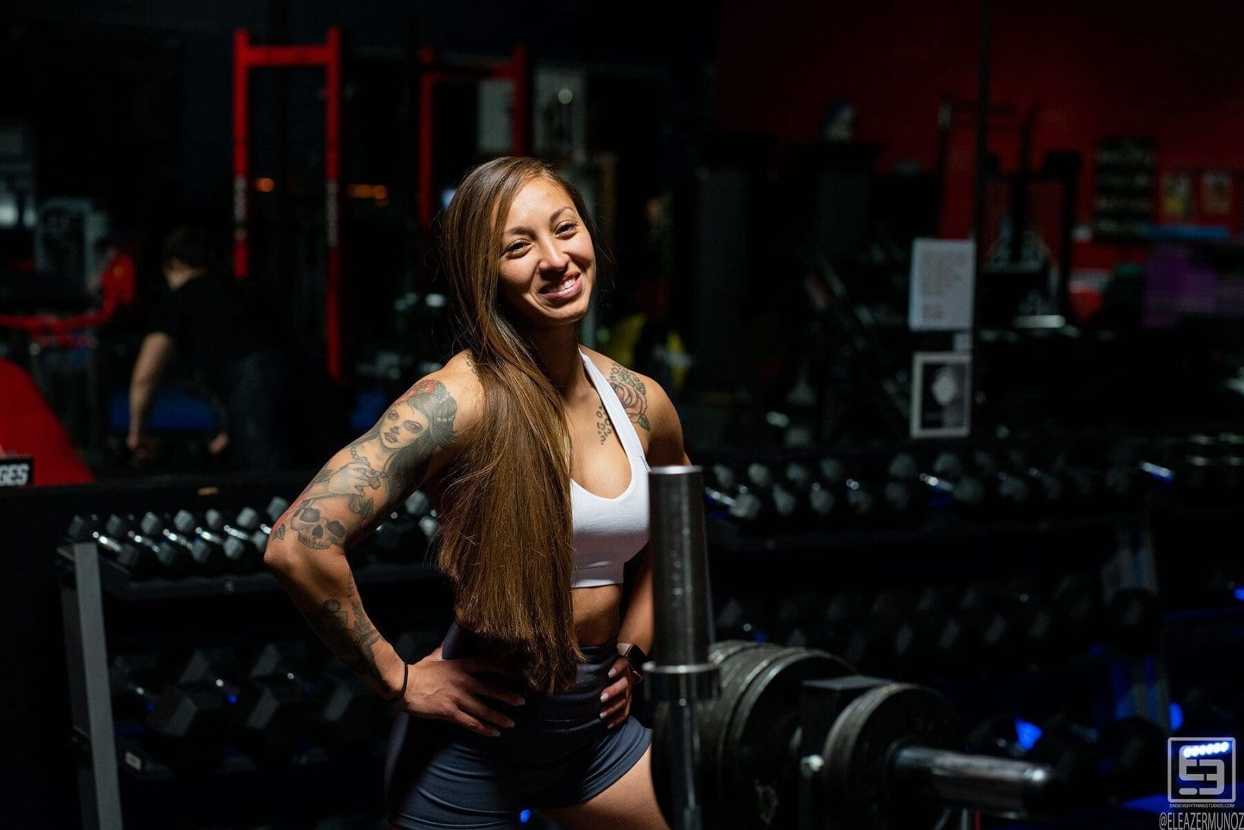 Danni stands with one hand on her hip and smiles beside the gym's weight area.