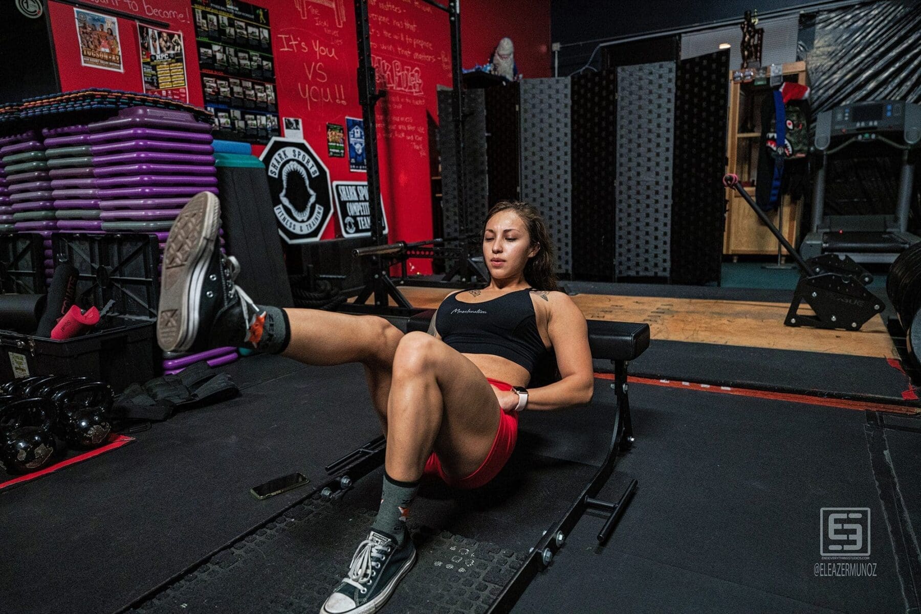 Danni leans against a low training bench while extending one leg inside a red-and-black gym.