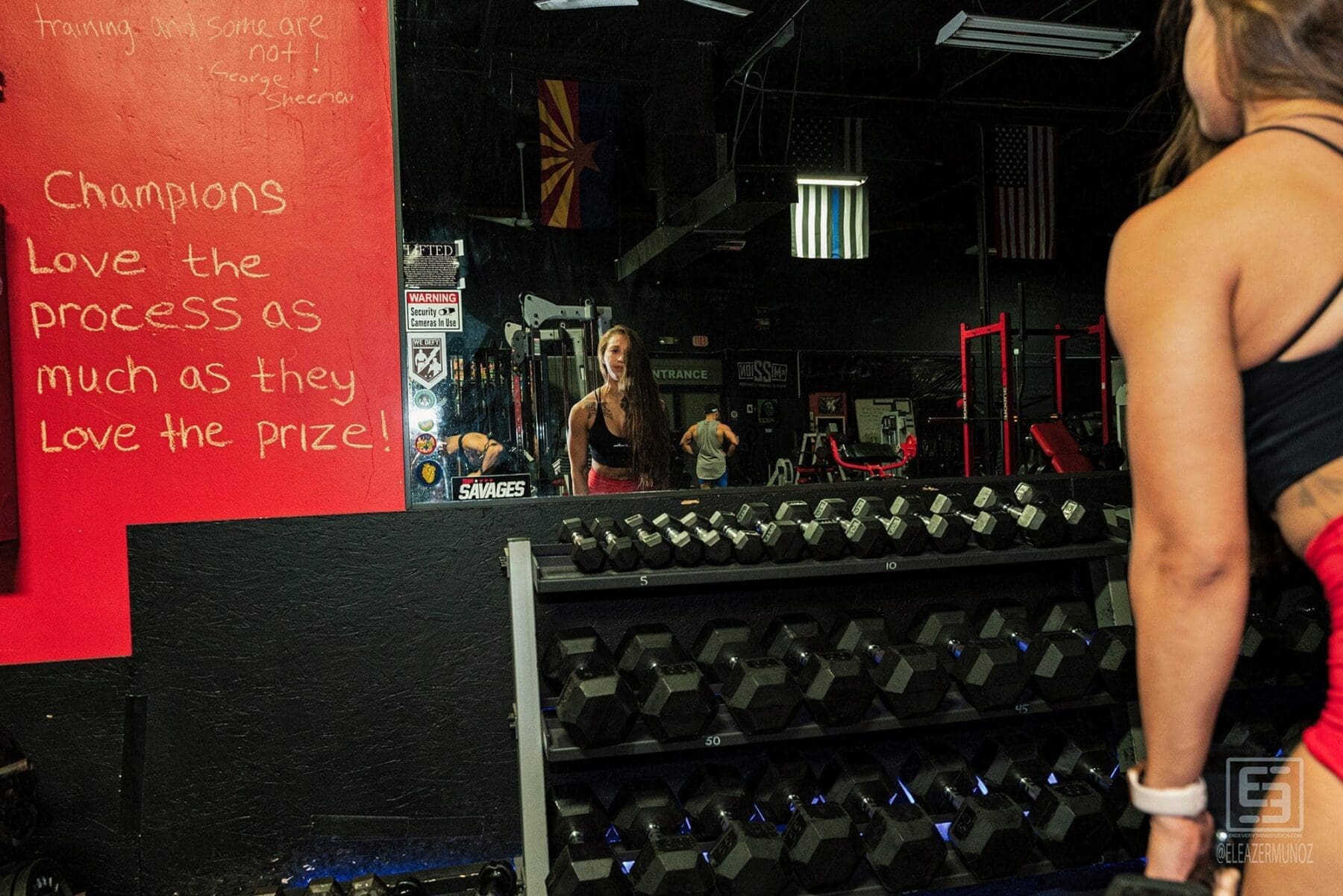 Danni holds a dumbbell in the foreground while her face and the gym appear in the wall mirror.