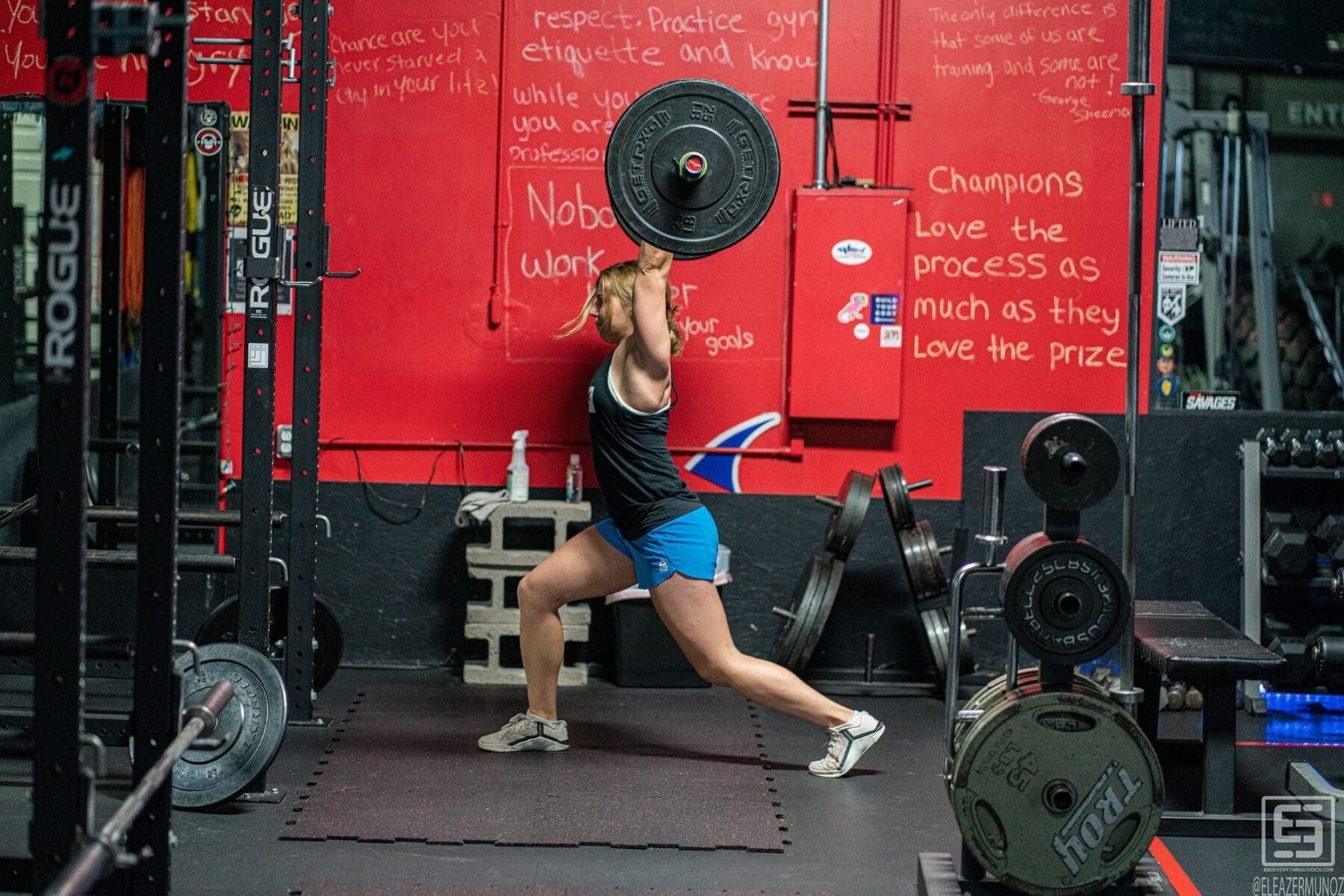 Day holds a weight plate overhead while stepping into a deep lunge beside a squat rack.