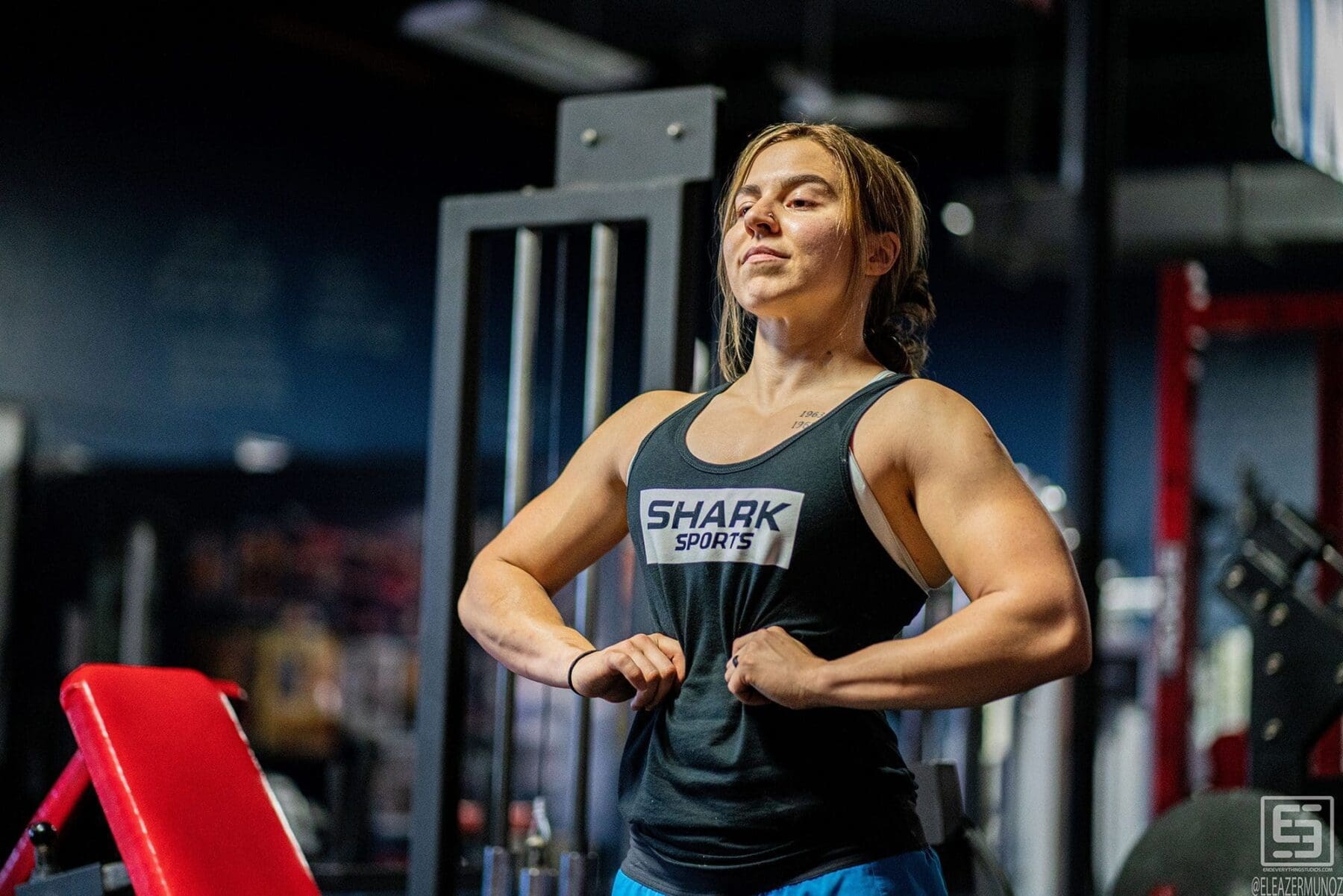 Day smiles slightly while holding a front upper-body pose beside a red training bench.