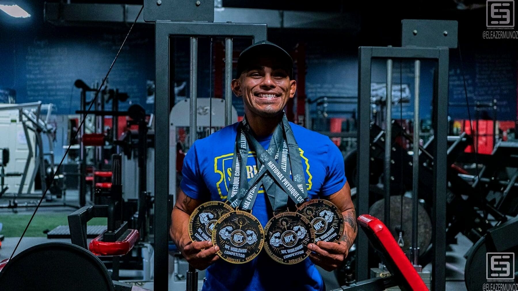 Christopher smiles in a blue shirt and black cap while holding several medals across his chest inside the gym.