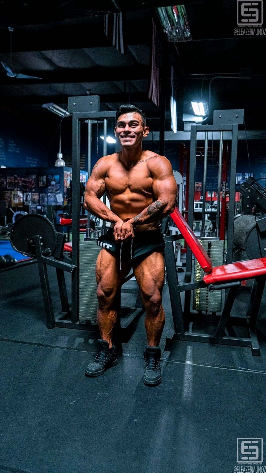 Christopher stands shirtless in a vertical full-body gym portrait and holds a front physique pose.