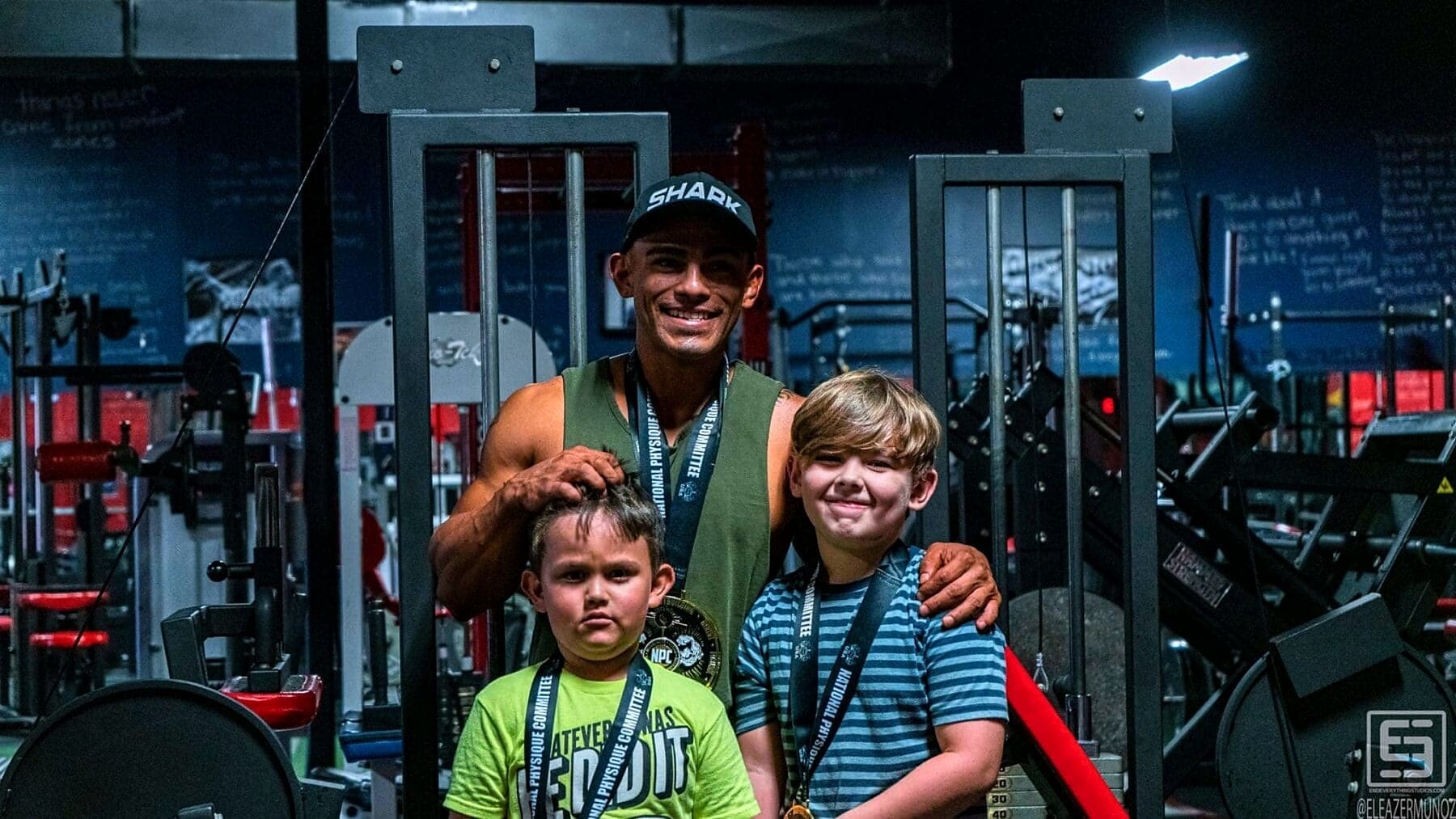 Christopher smiles beside two children as all three display medals inside the gym.