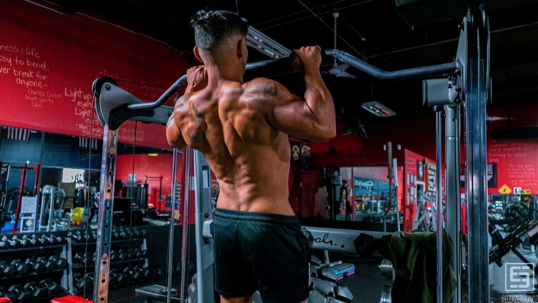 Christopher performs a pull-up from behind, revealing his back and shoulder definition against a red gym wall.