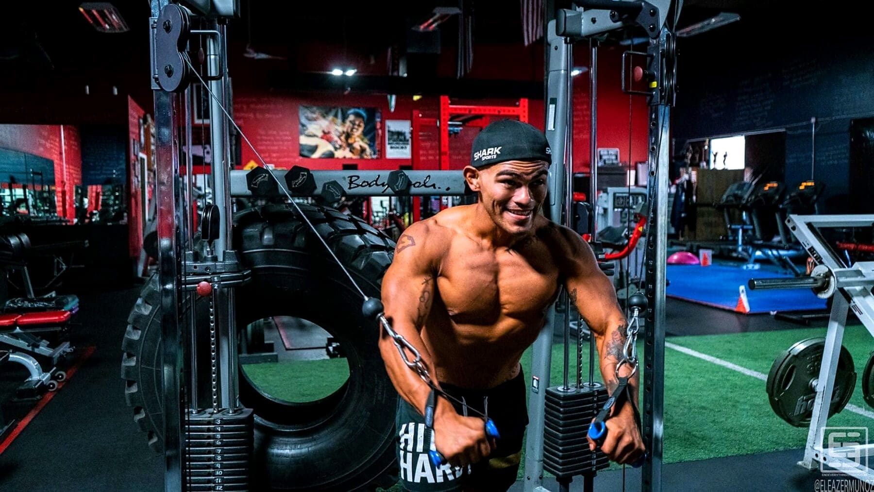 Christopher leans forward between two cable stacks and draws blue handles inward during a standing chest exercise.