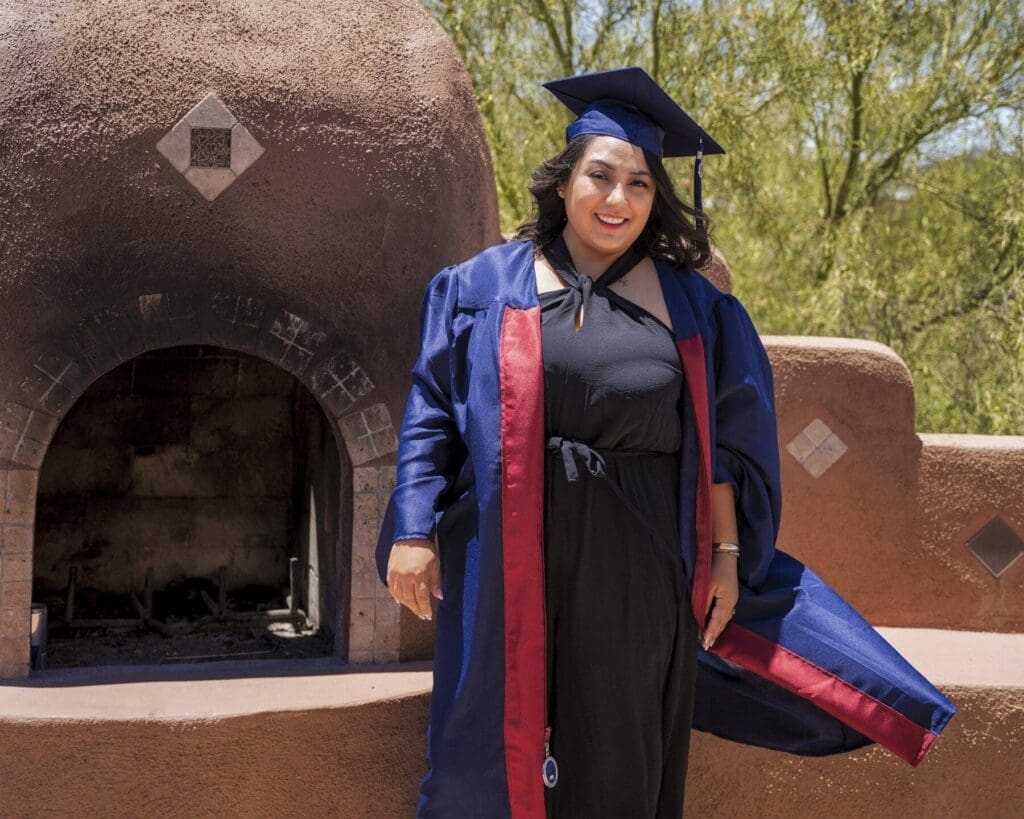 Hilda wears her blue graduation cap and gown while standing beside a rounded outdoor fireplace.