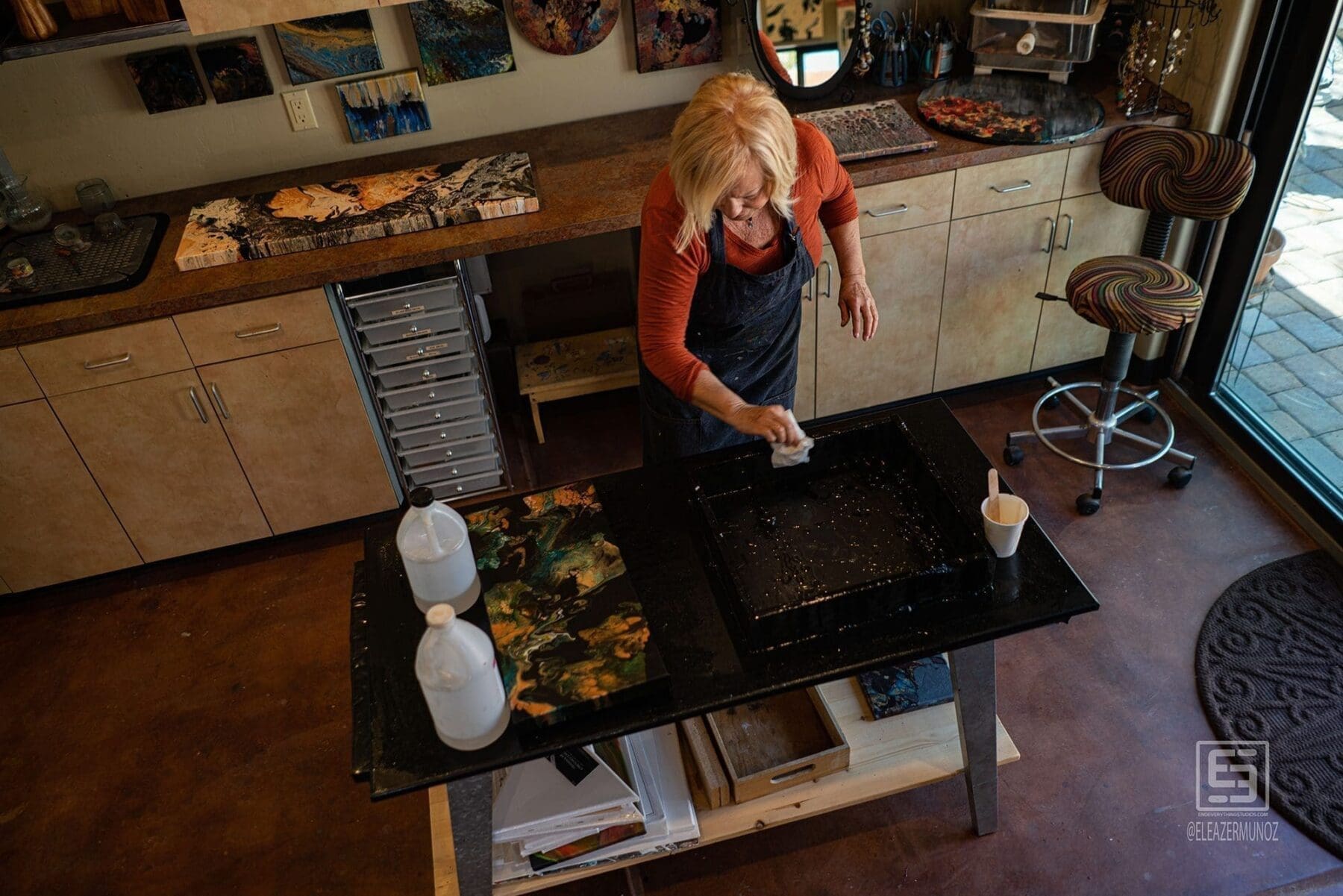 An overhead view shows the artist working at a central studio table surrounded by artwork, tools, and materials.