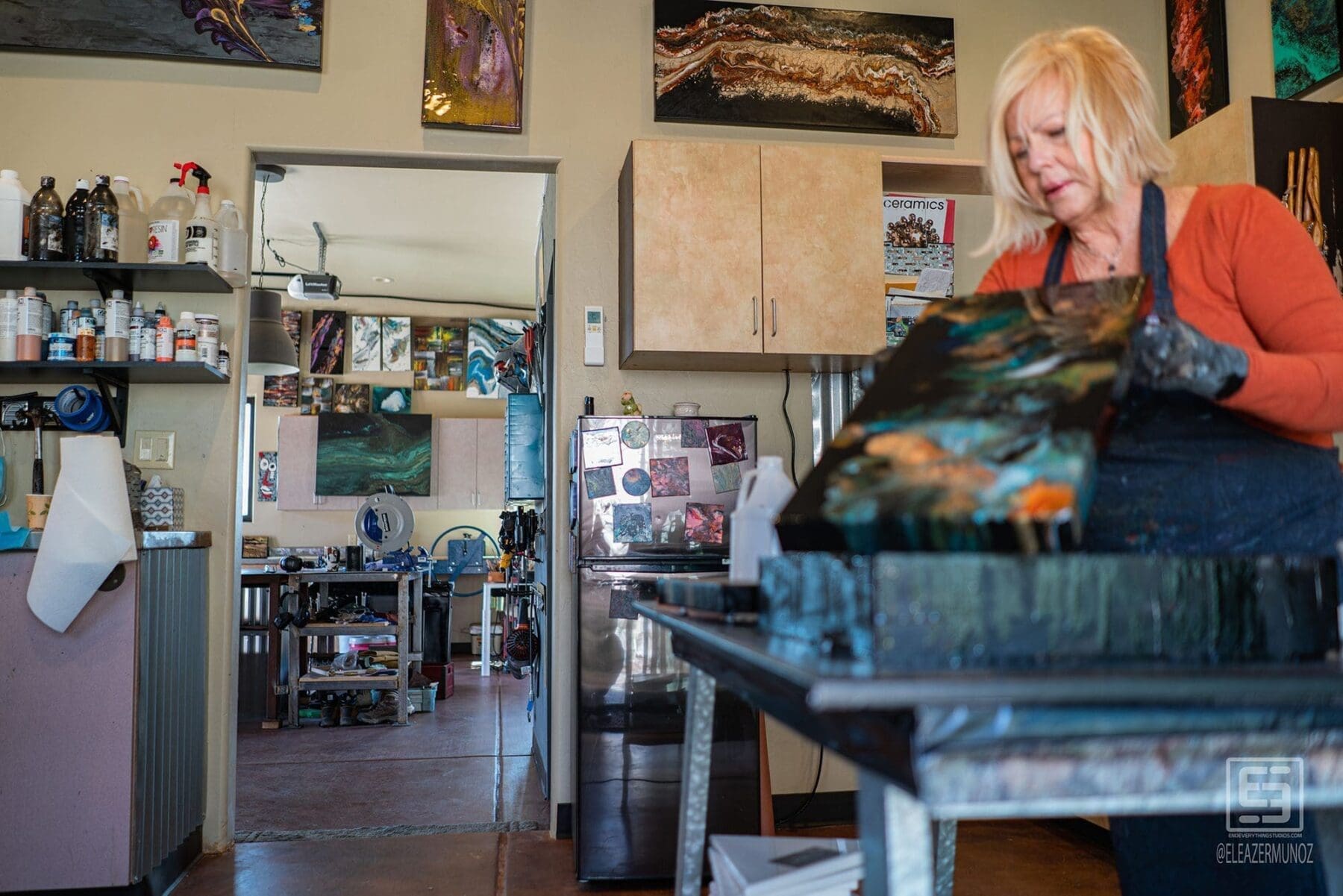 A low close view follows a glossy, colorful artwork toward the artist working at the far end of the table.
