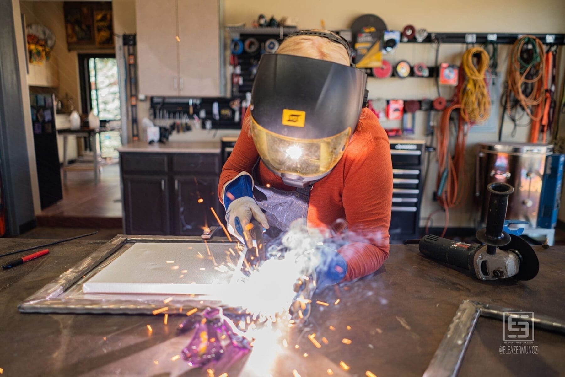 A brilliant burst of sparks fills the foreground as the artist welds a metal frame at the studio workbench.