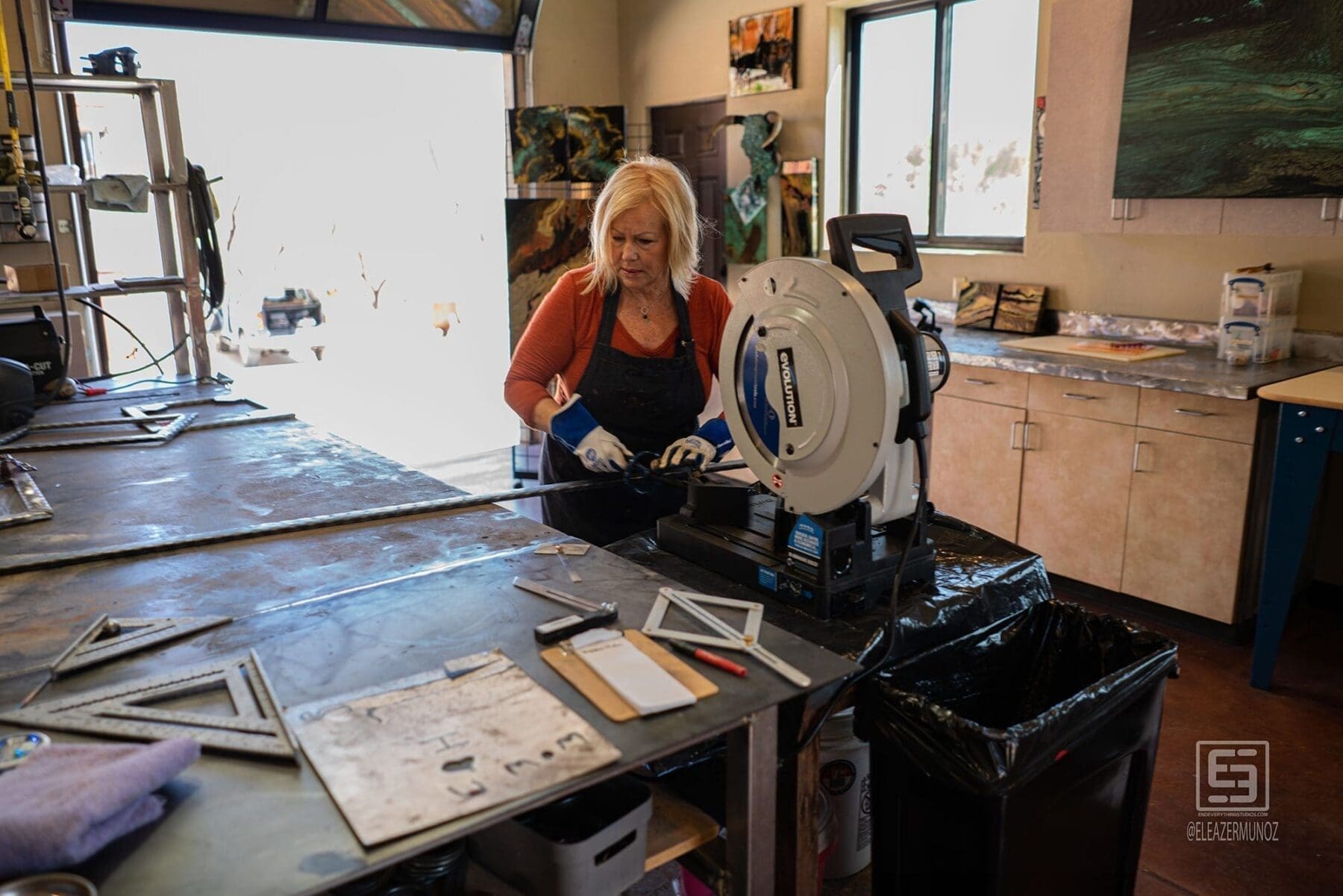 The artist operates a chop saw at the workbench with cut frame pieces and measuring tools nearby.