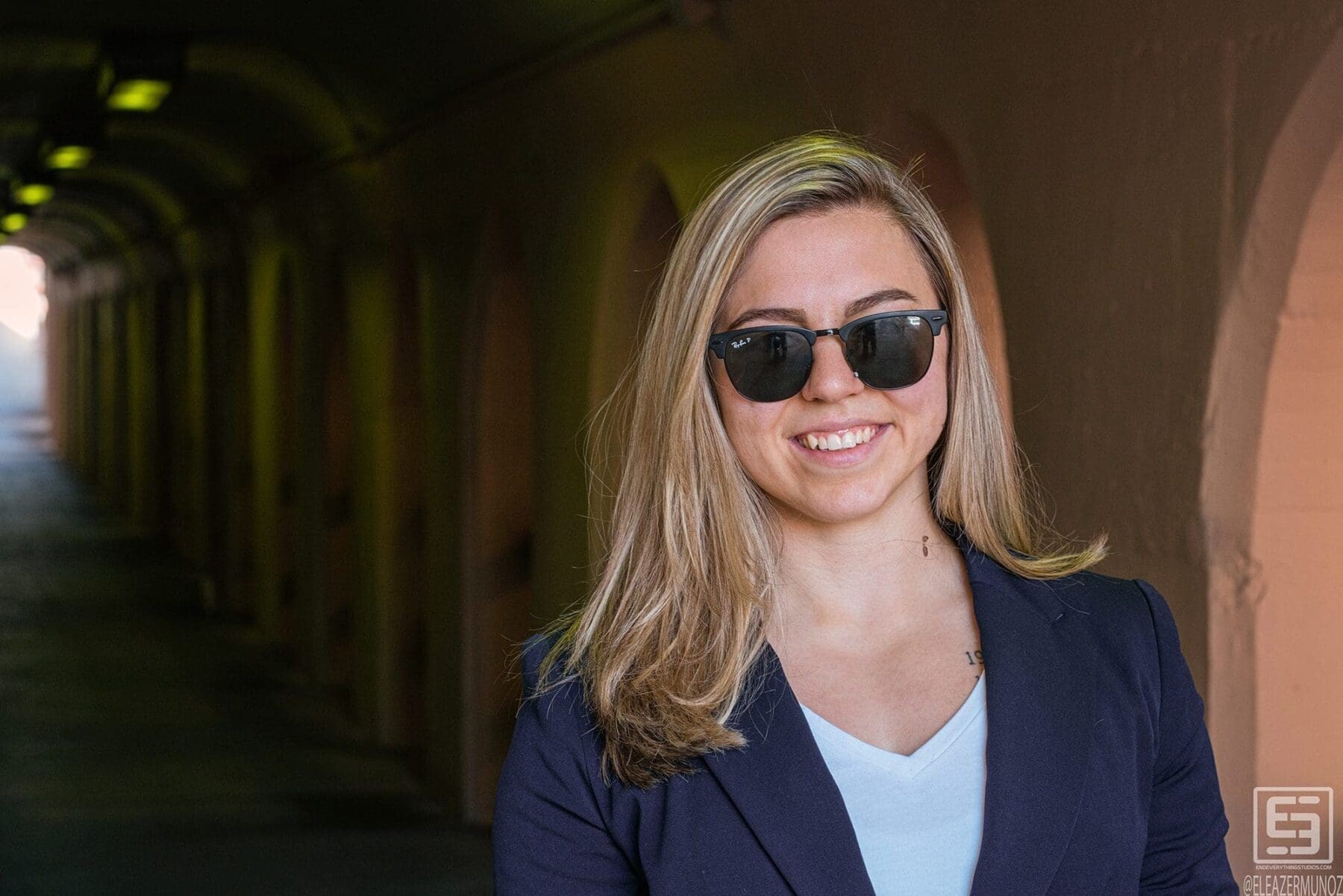 Day Muñoz smiles in a navy blazer and sunglasses with a long arched passage receding behind her.