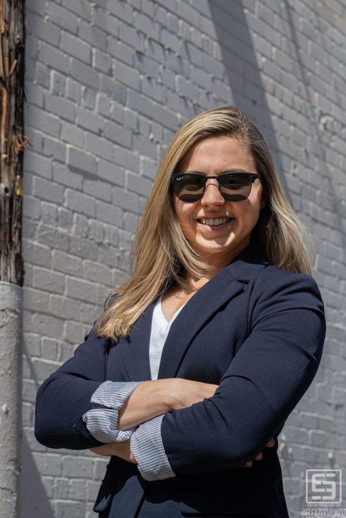 Day Muñoz stands with arms crossed in a navy blazer and sunglasses against a dark painted brick wall.