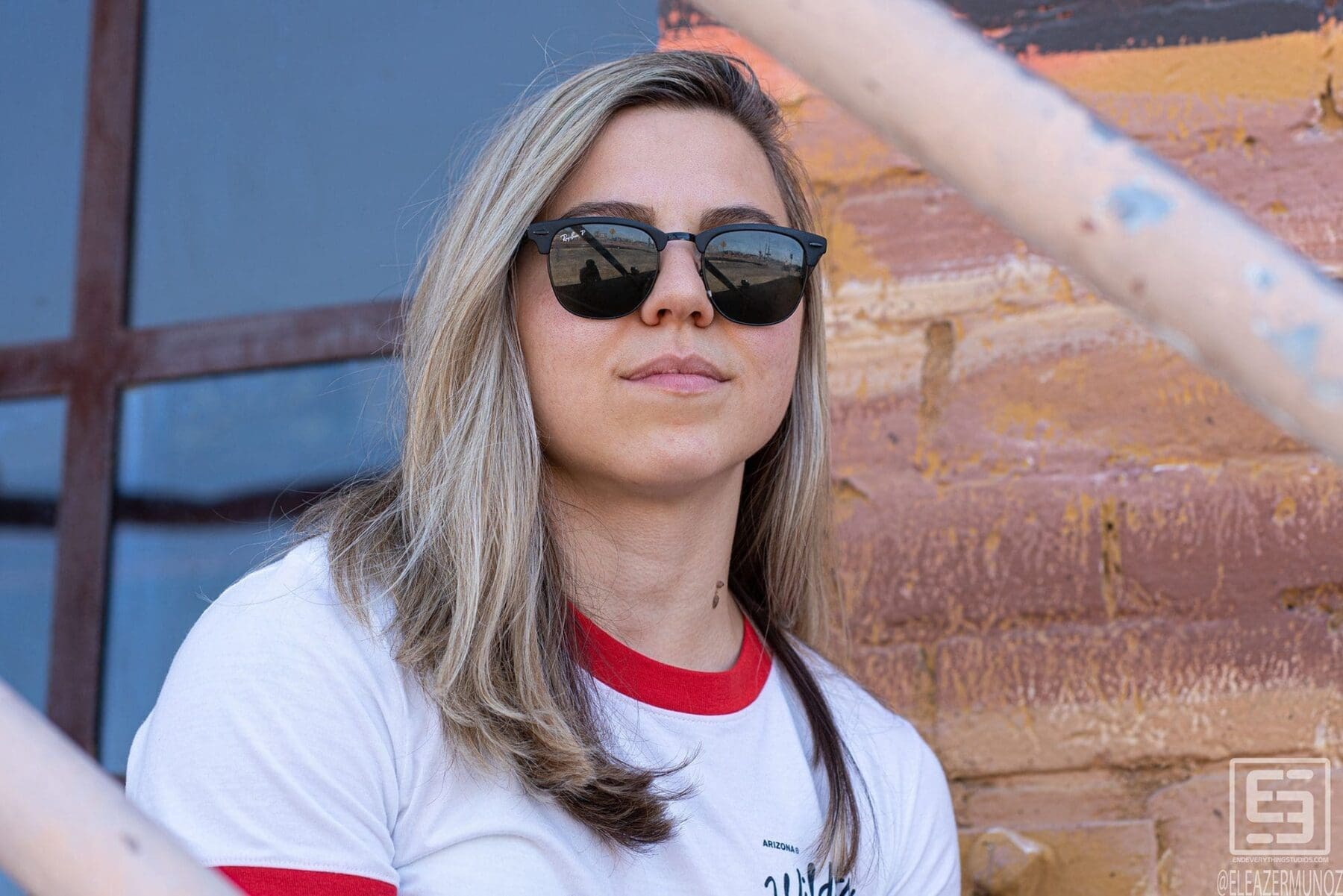 Day Muñoz wears sunglasses and a white ringer T-shirt for a close portrait beside colorful painted brick and metal rails.