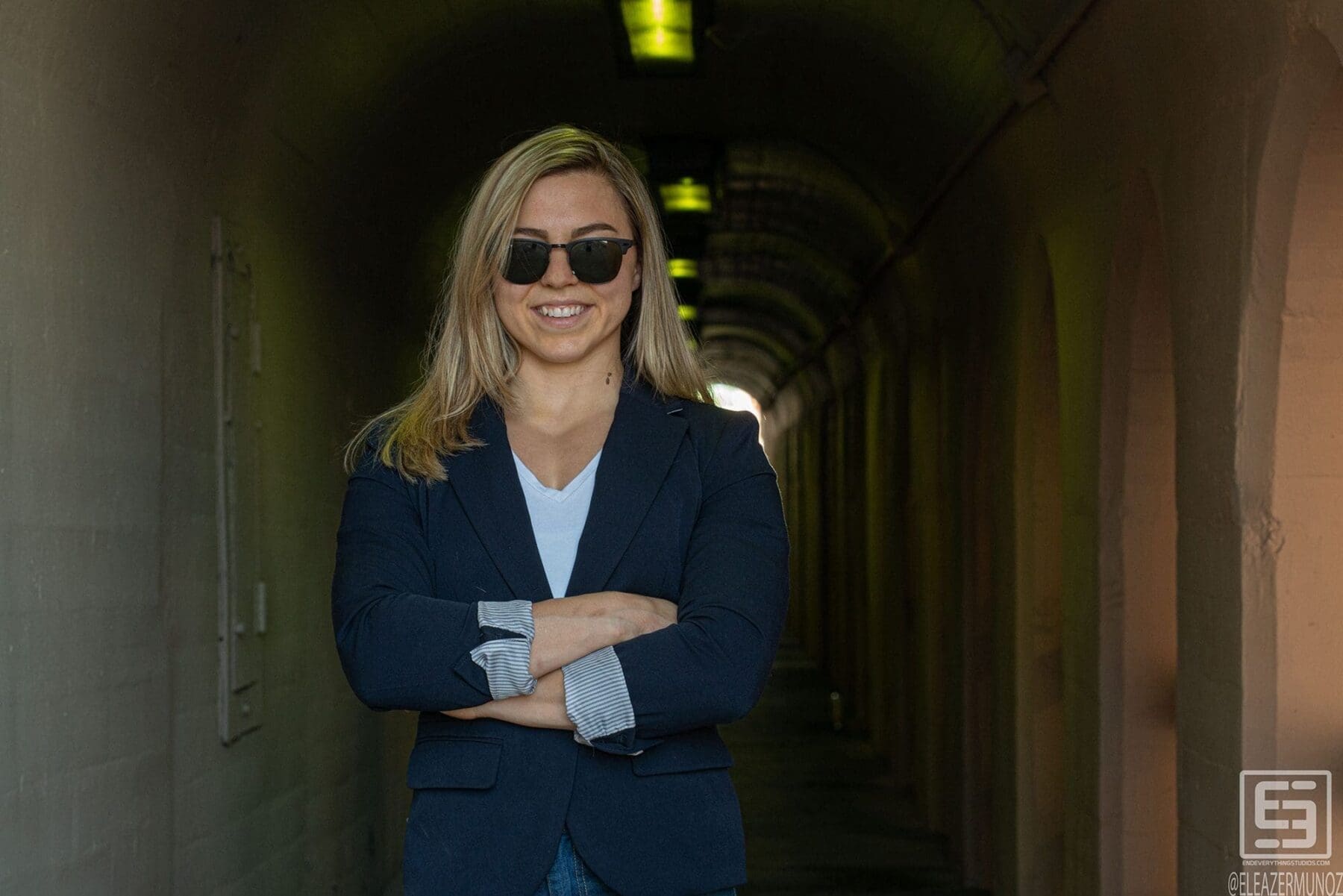 Day Muñoz smiles with arms crossed in a navy blazer as the arched corridor recedes behind her.