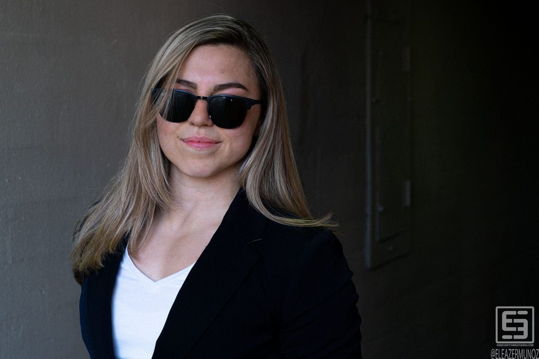 Day Muñoz smiles in sunglasses and a navy blazer against a dark shaded wall.
