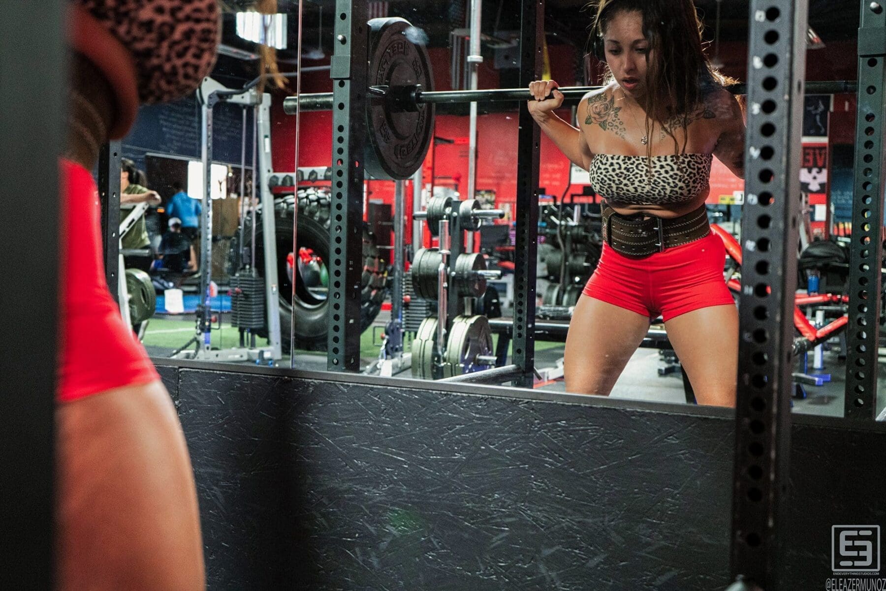 Danni squats under a barbell while facing her reflection in the gym mirror.