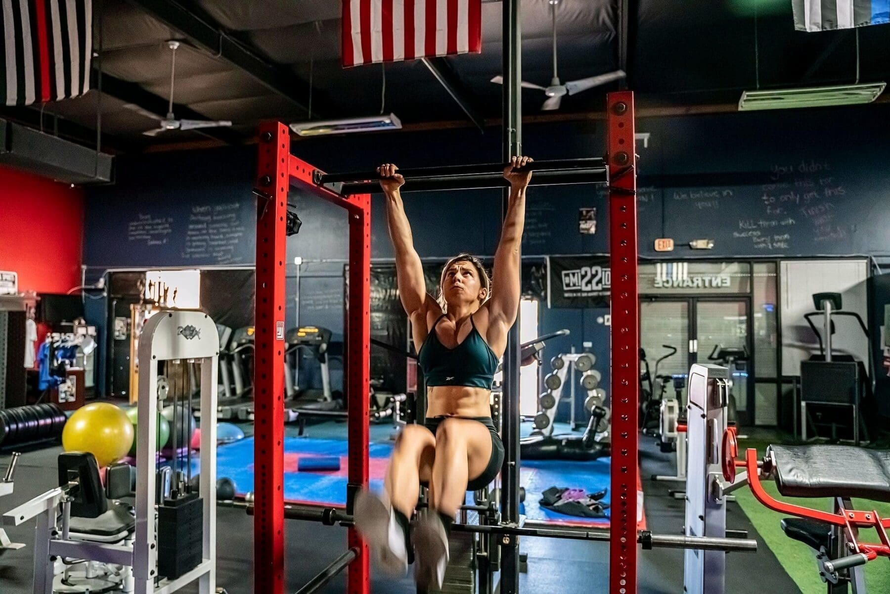 Day hangs from a pull-up bar with one leg extended forward inside a red rack.