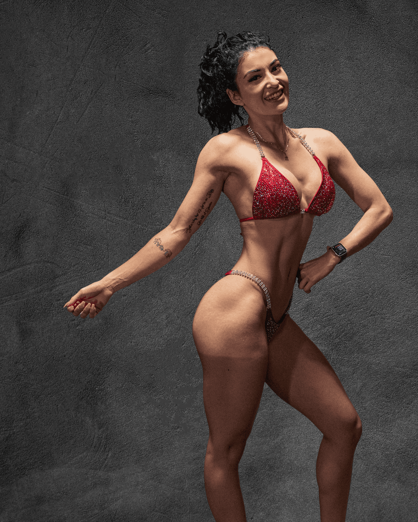 Cynthia smiles in a side physique pose wearing a red rhinestone bikini.