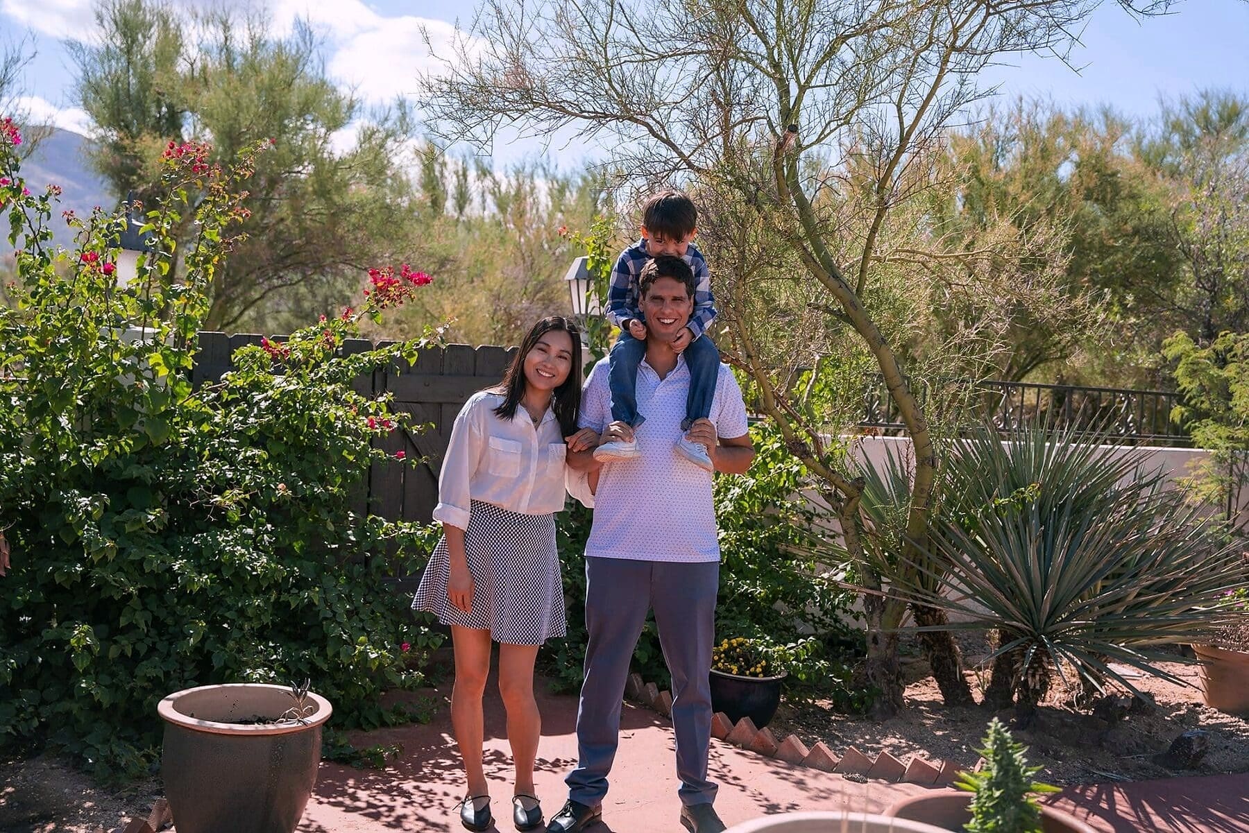 Three family members smile in a garden while a child rides on an adult's shoulders.