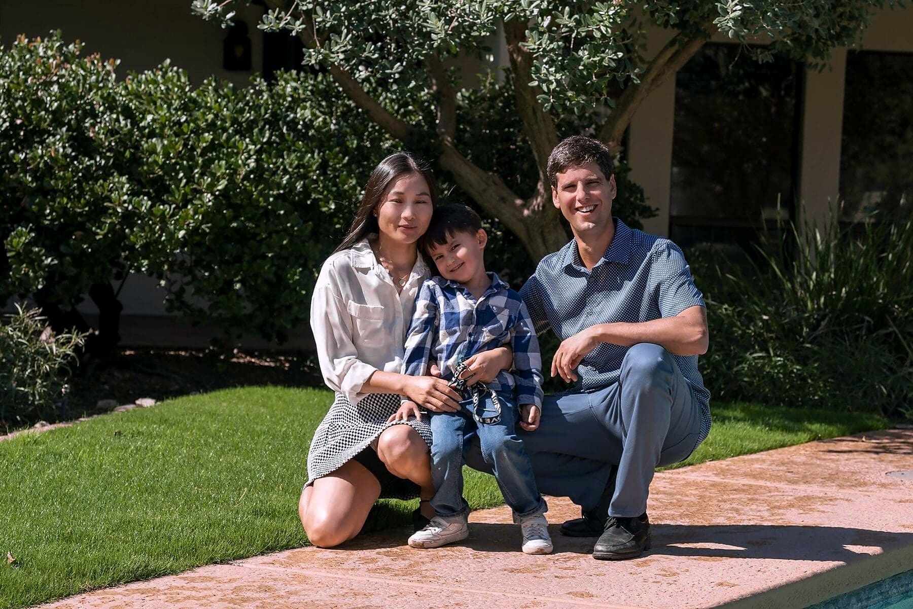 Three family members sit closely together on a sunlit patio beside a swimming pool.