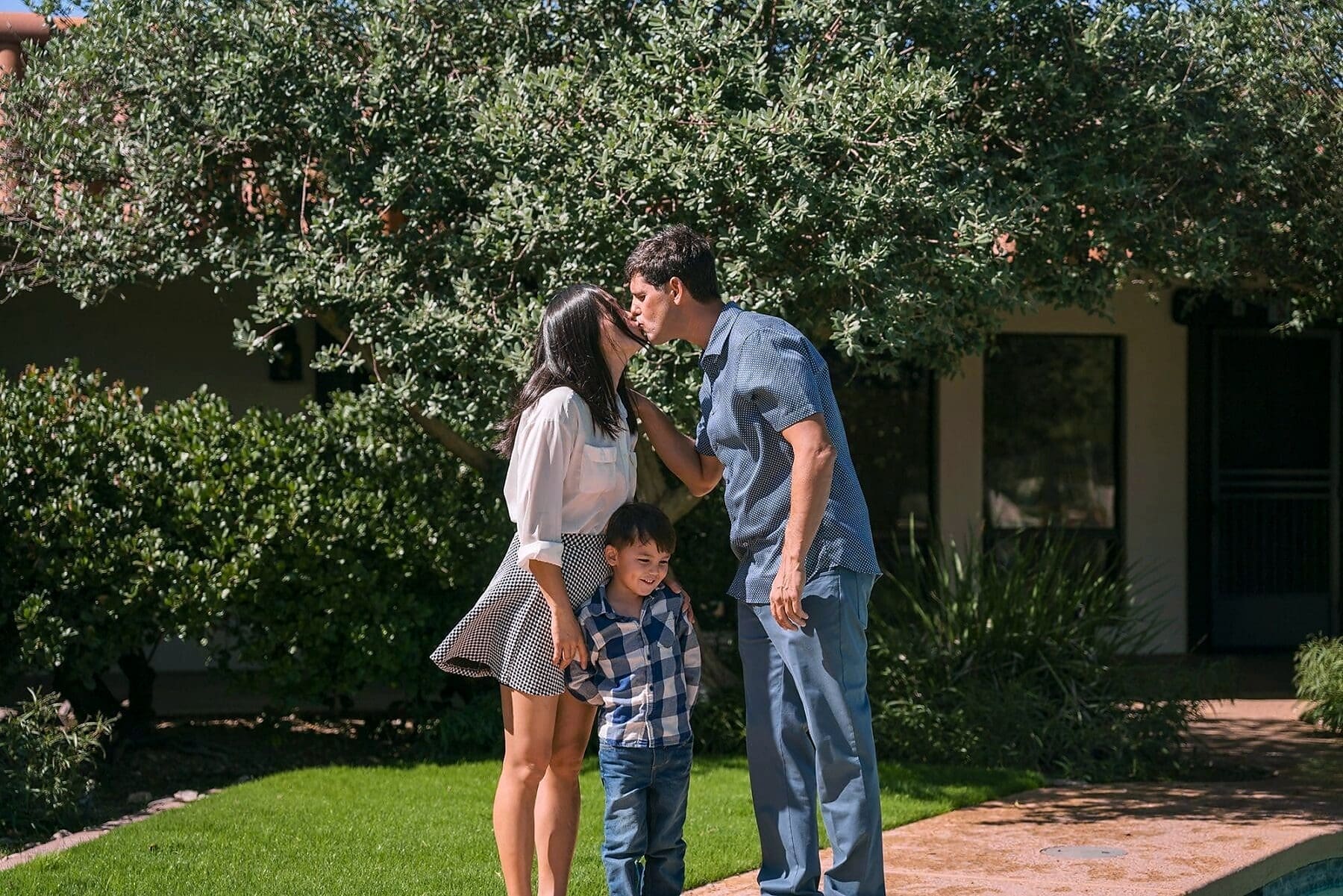 Two adults share a kiss while a child stands between them on a sunlit lawn.