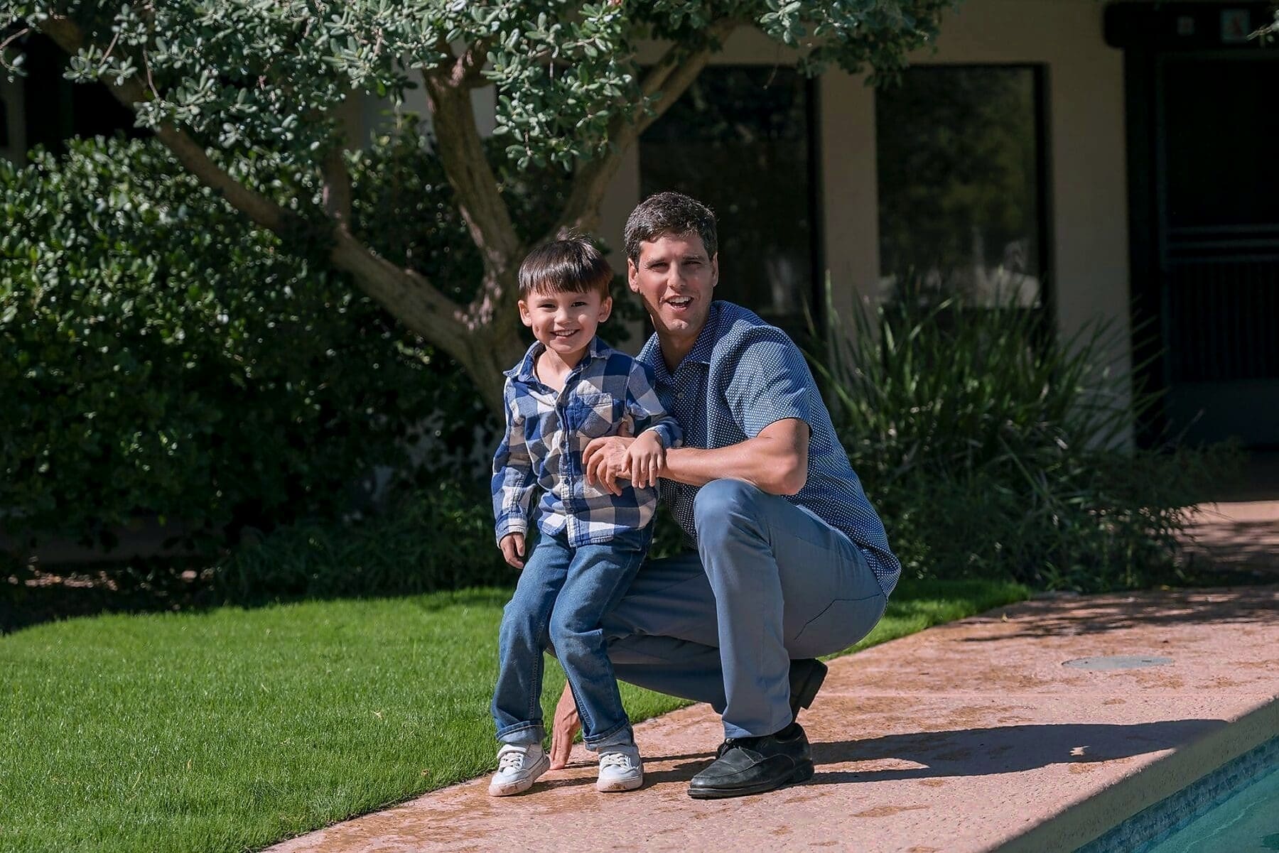 An adult and child crouch together on a stone patio beside a swimming pool.