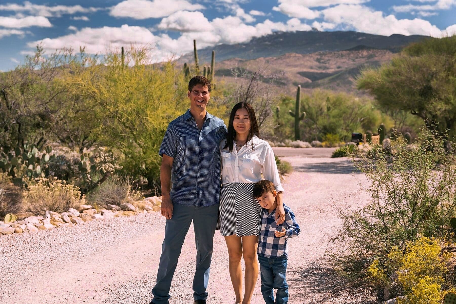 Three family members stand together on a desert path with cacti, flowering shrubs, and mountains behind them.