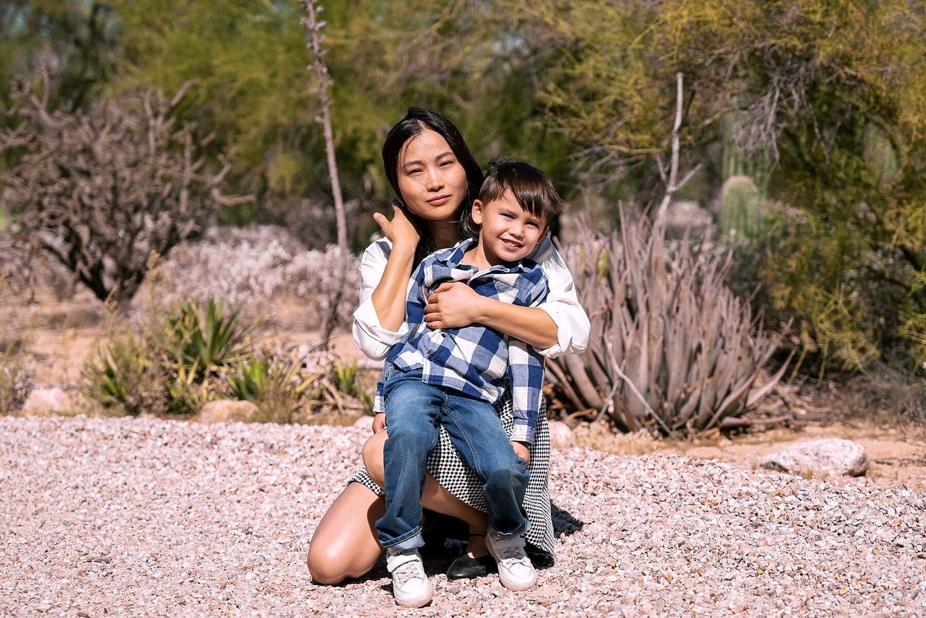 An adult kneels on gravel and holds a smiling child against a backdrop of desert plants.