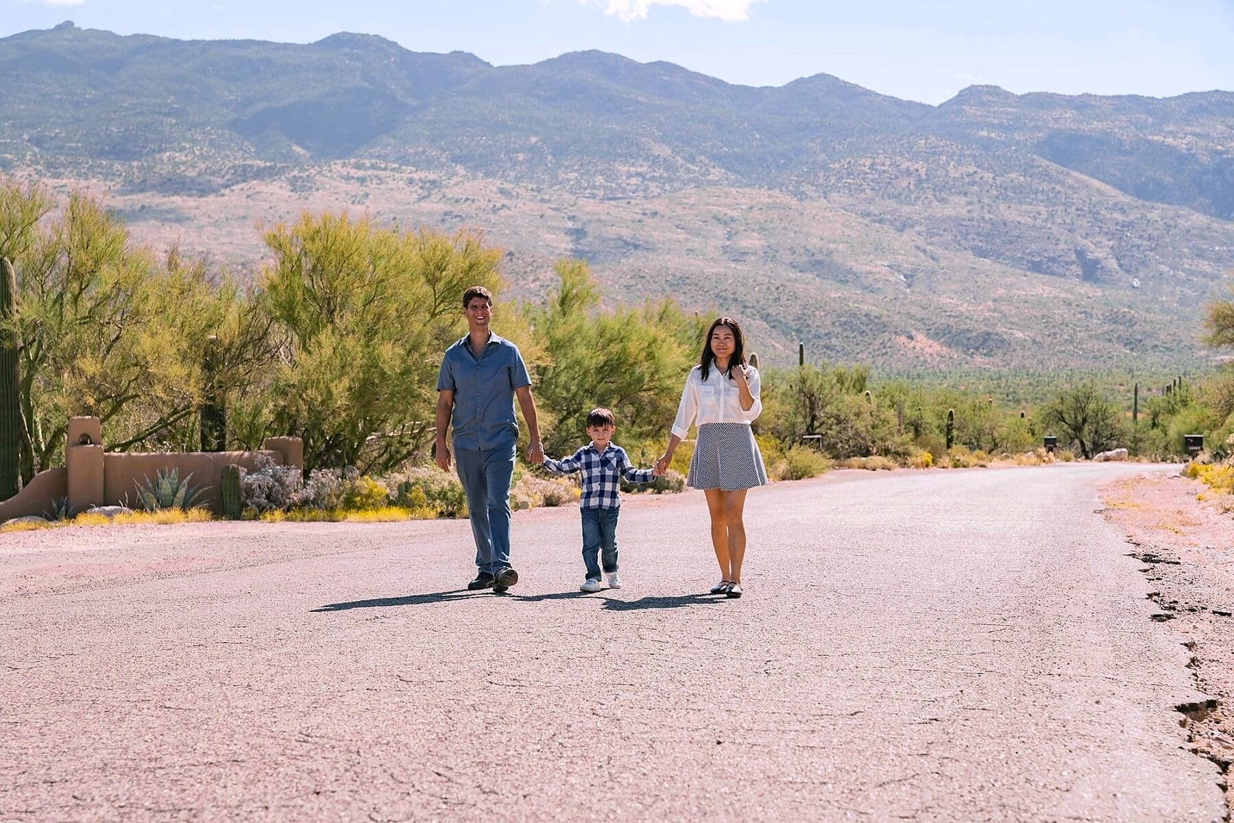 Three family members walk hand in hand on a sunlit road with a broad mountain ridge behind them.