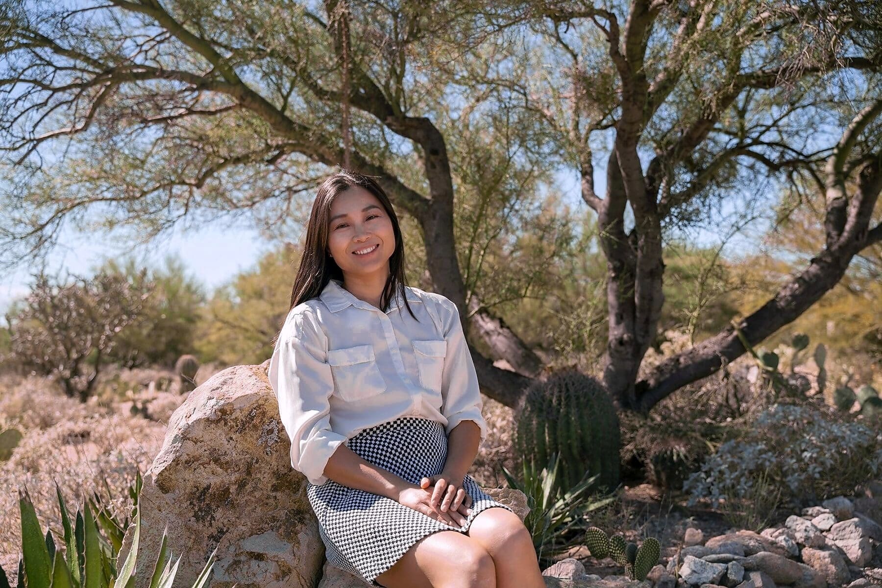 An adult sits smiling on a pale rock beneath the spreading branches of a desert tree.