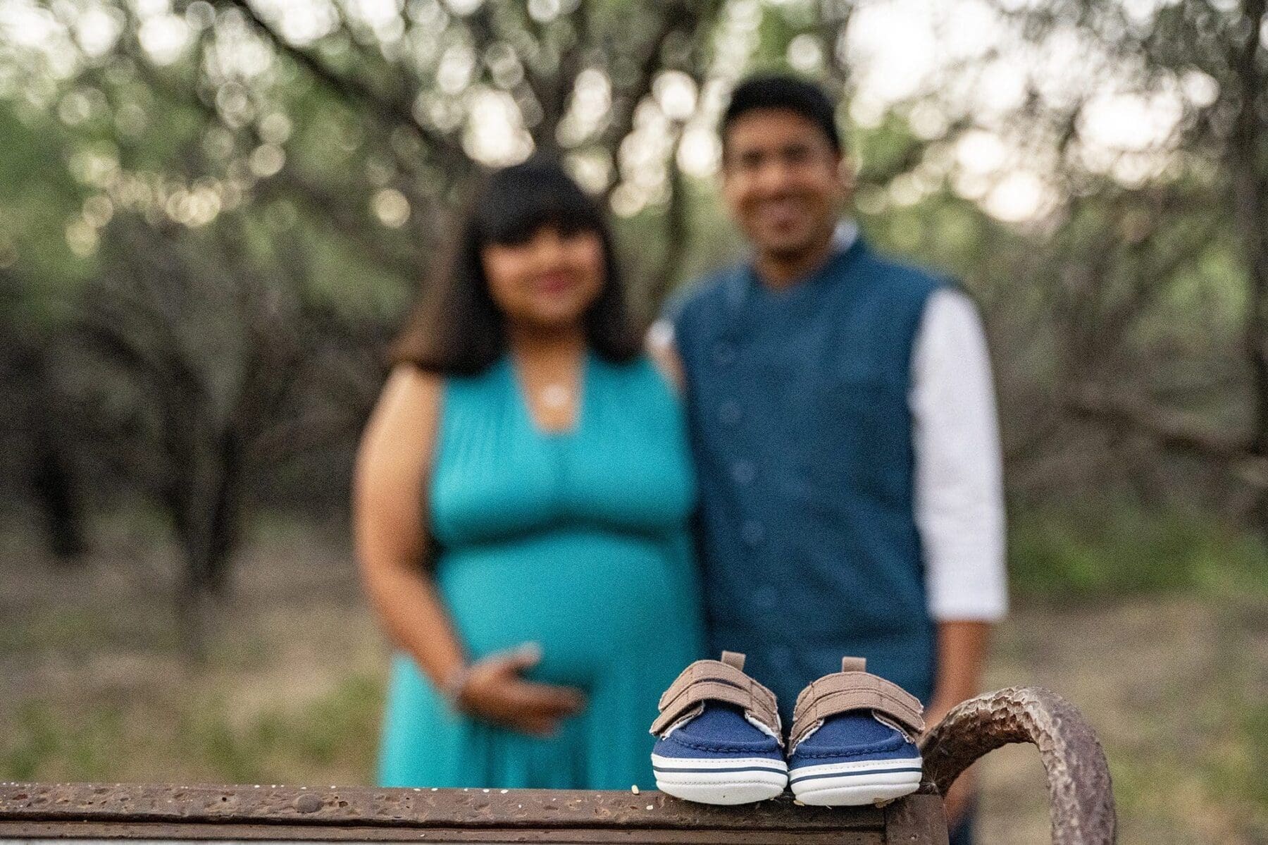 A pair of baby shoes rests in sharp focus while Aditi and her partner stand softly blurred in the wooded background.