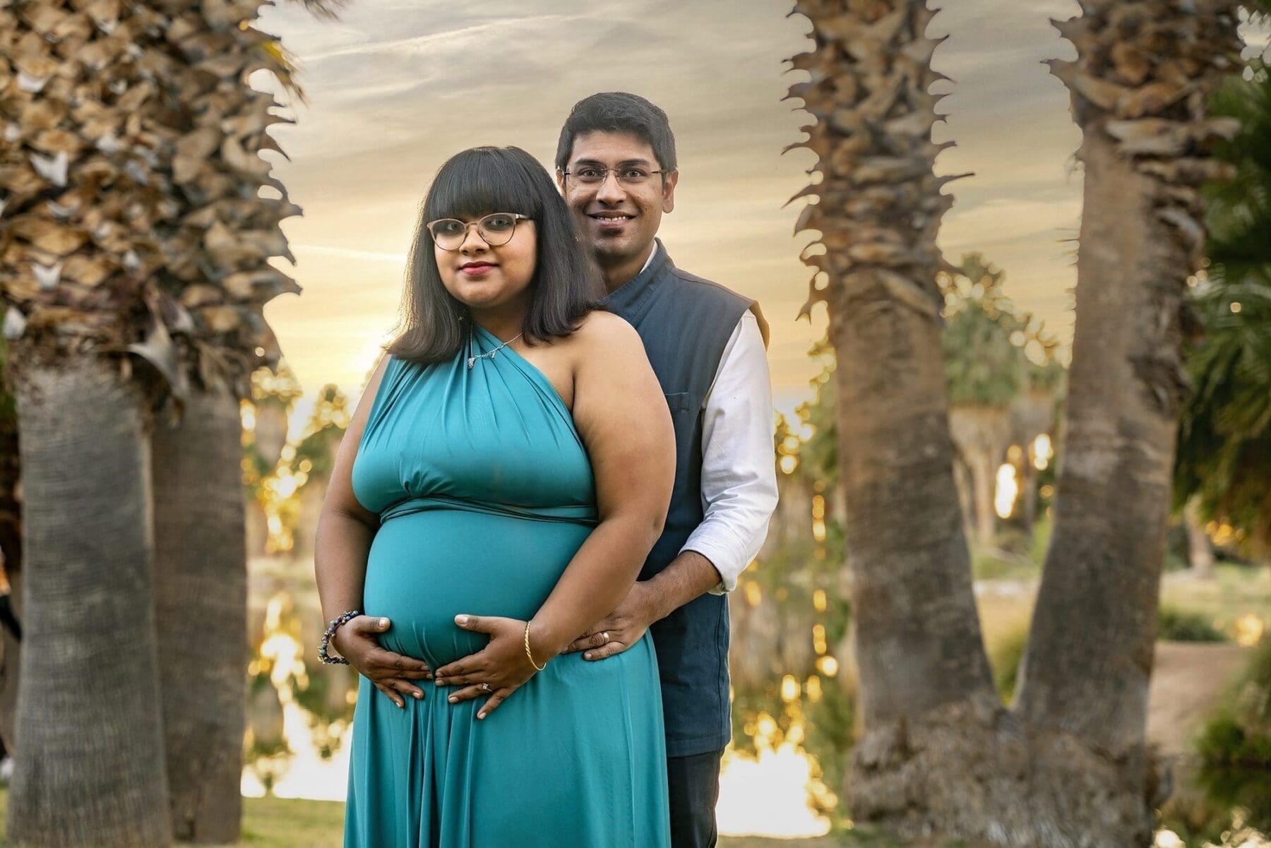 Aditi faces the camera with hands resting on her belly while her partner stands closely behind her among palm trees.