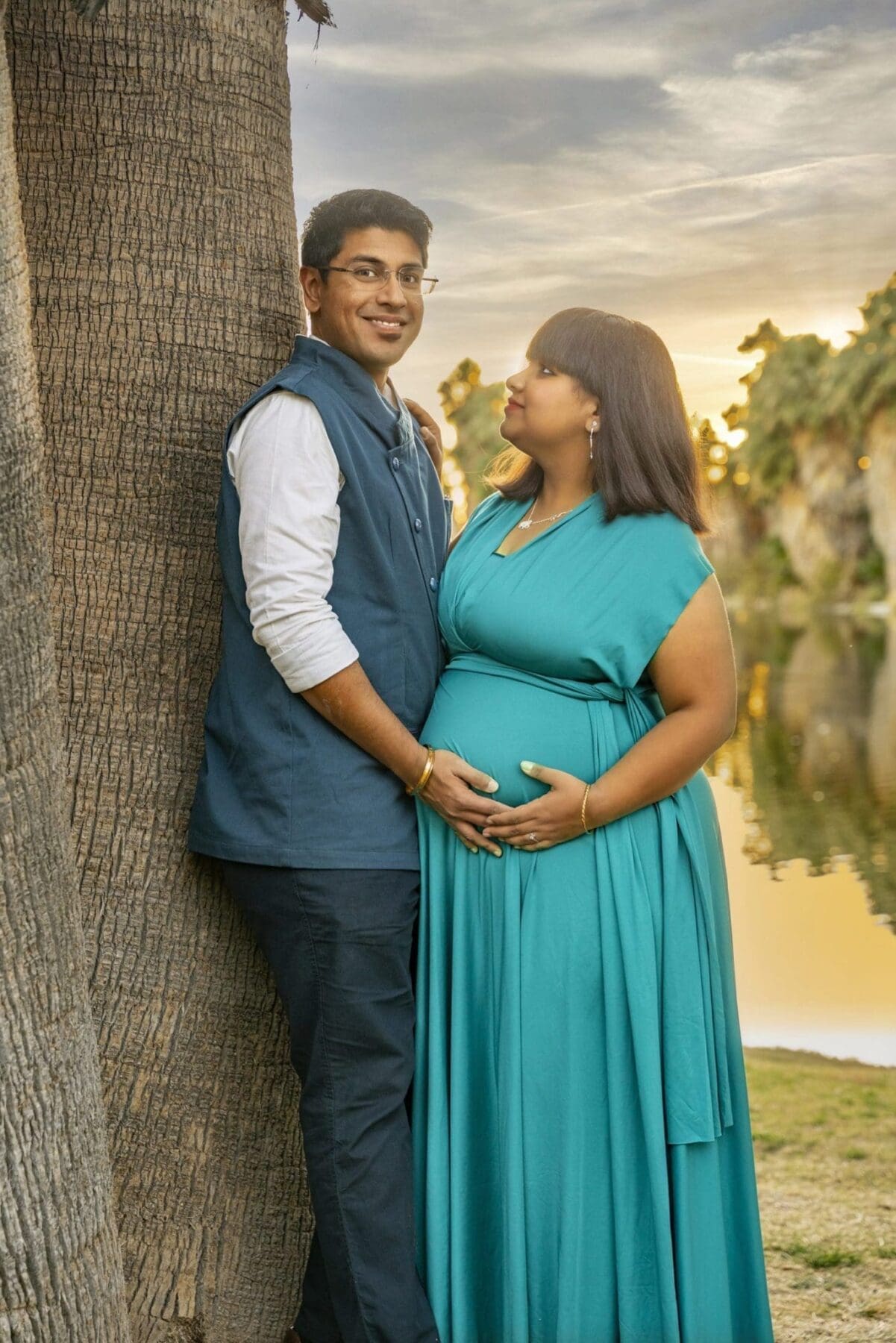 Aditi looks toward her partner while they stand closely together beside a palm trunk and pond.