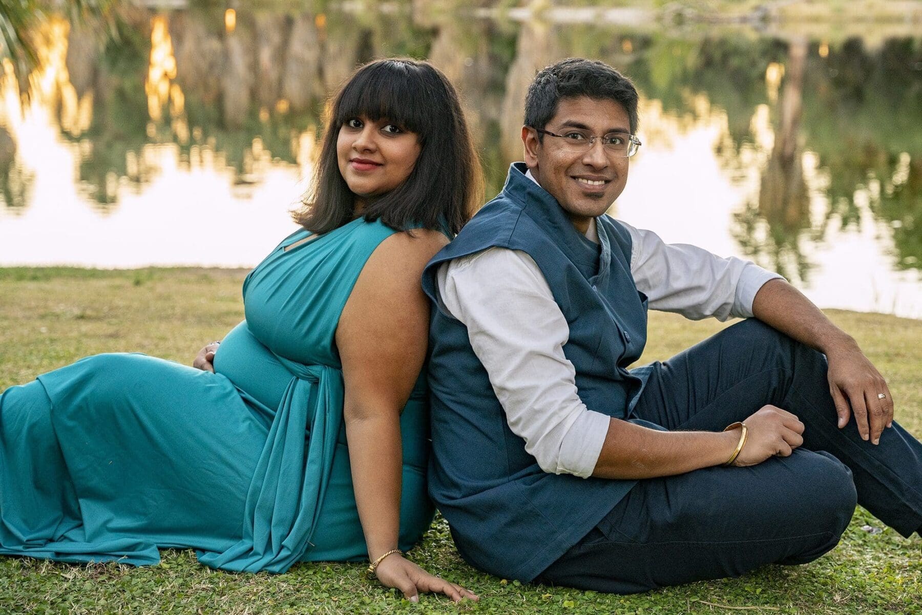 Aditi and her partner sit back to back on the grass beside the pond, both facing the camera.