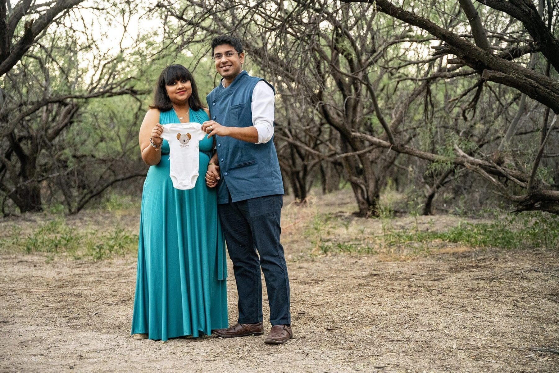 Aditi and her partner face the camera while holding up an infant bodysuit beneath desert trees.
