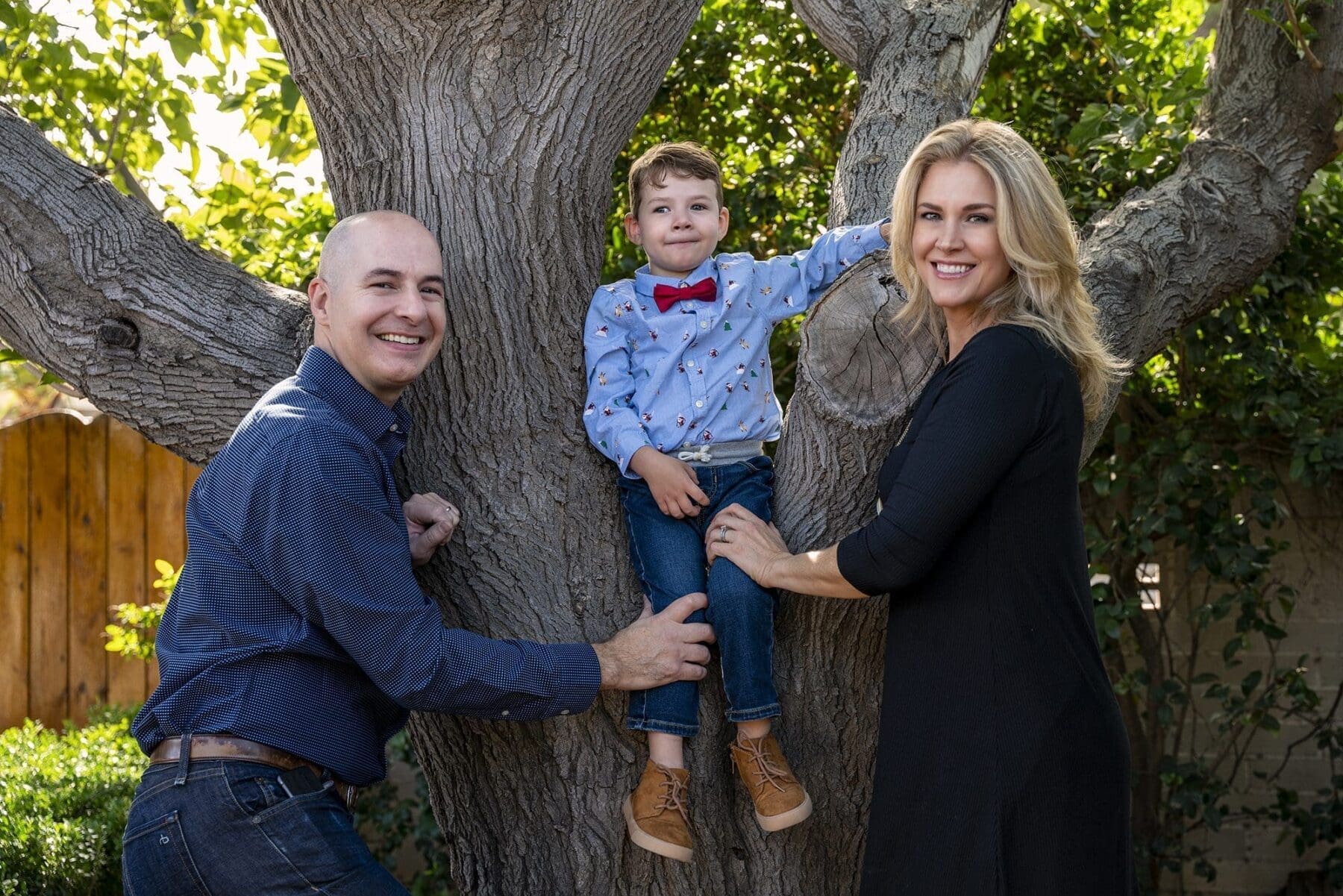 Two adults smile beside a young child standing against a broad tree trunk.