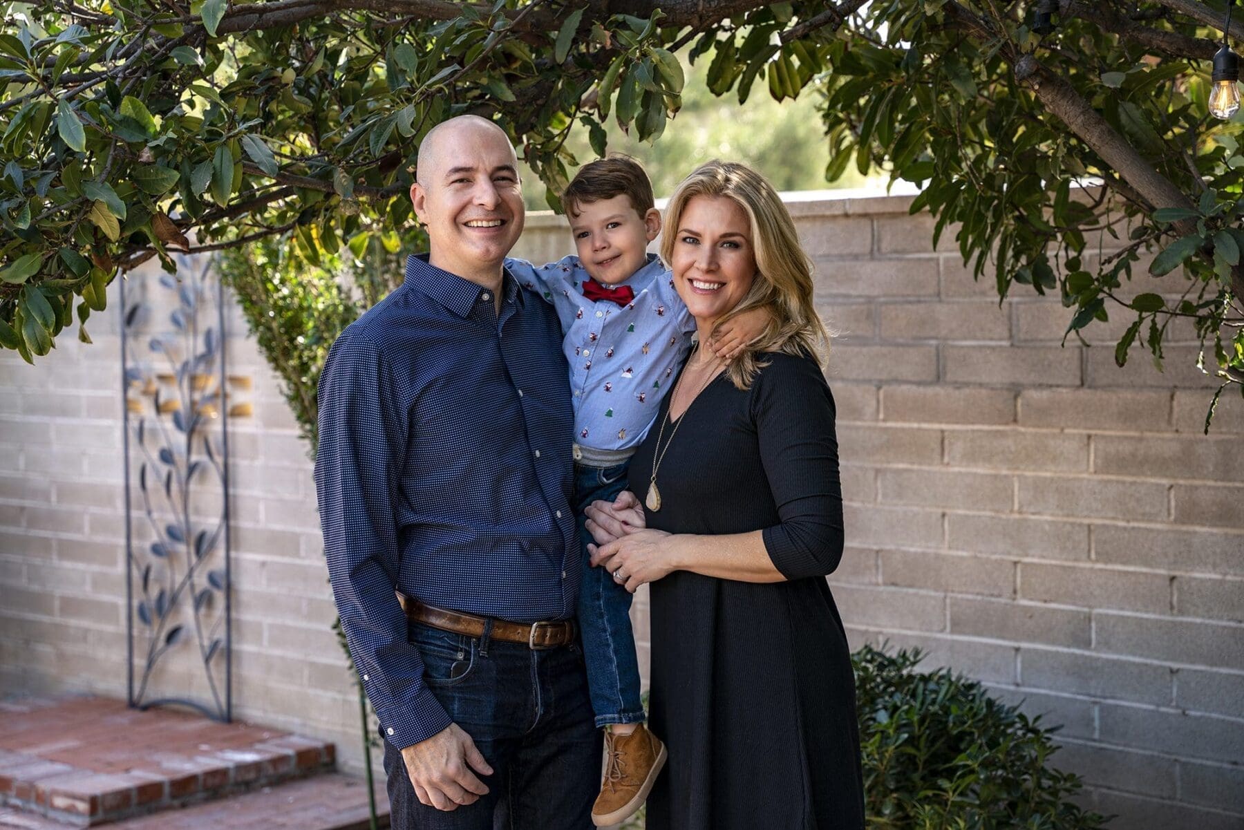 Two adults hold a young child in a close outdoor portrait beneath green branches.