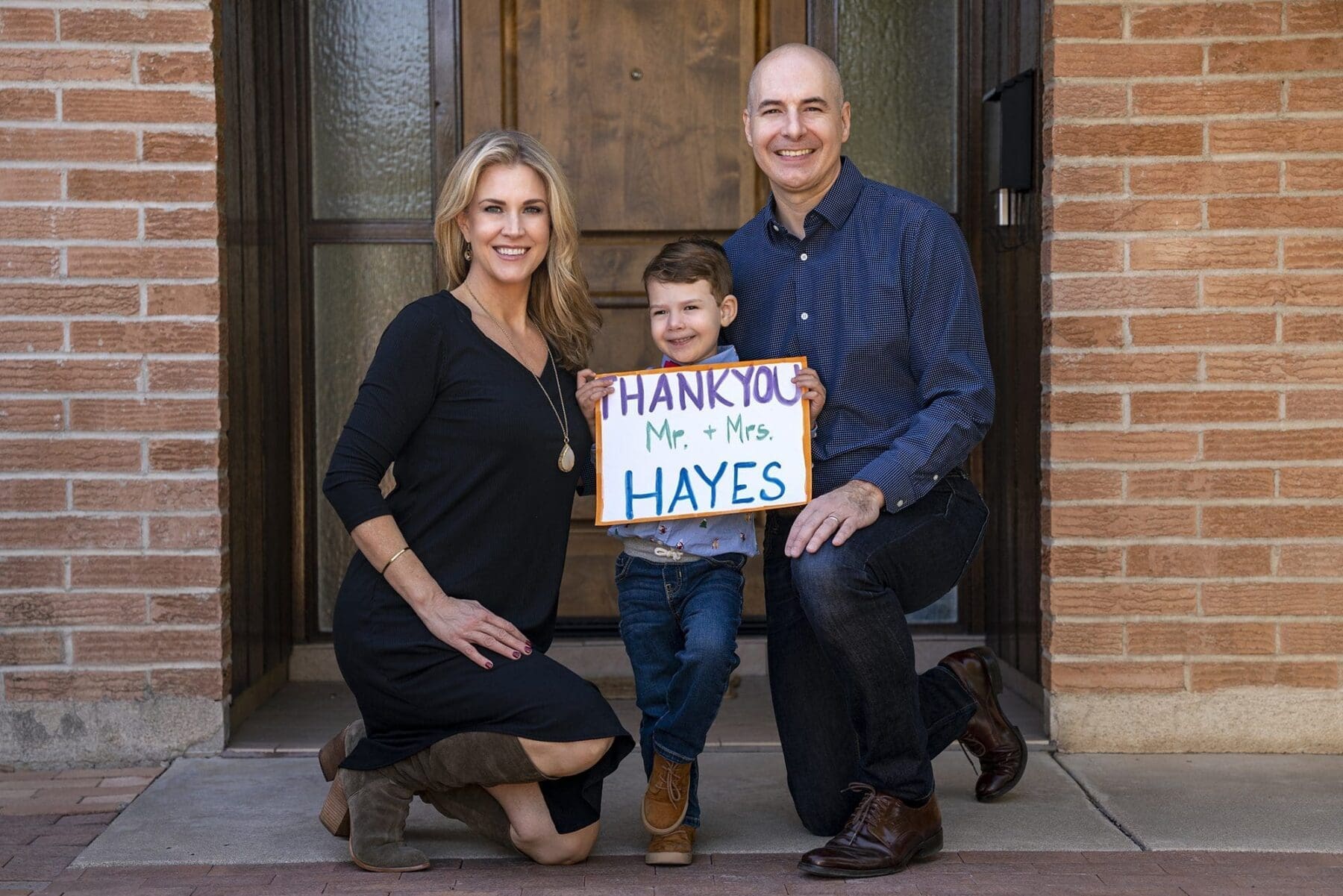 Two adults kneel beside a young child holding a hand-lettered thank-you sign at a brick entryway.