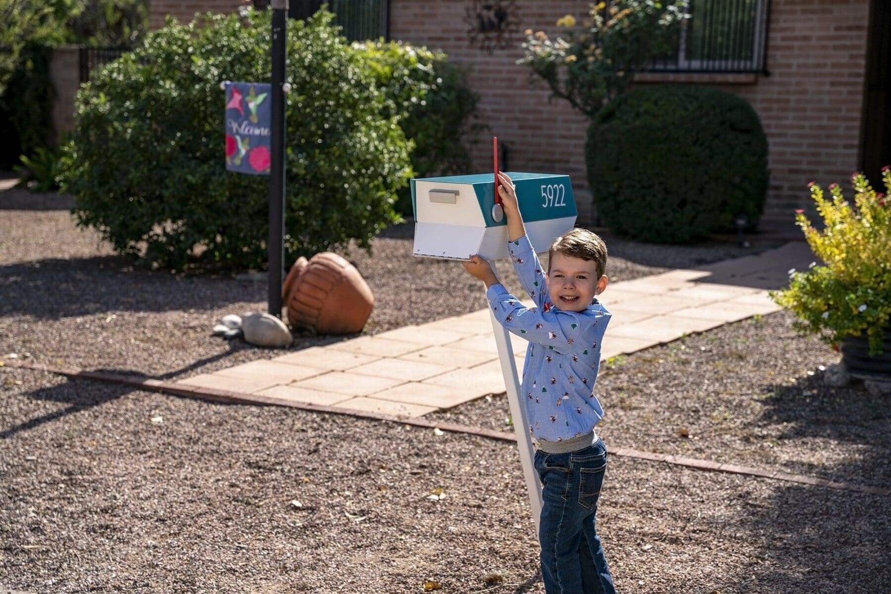 A young child walks across a gravel yard while lifting two colorful rectangular boxes.