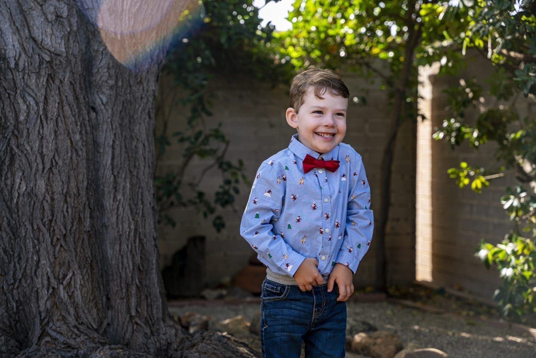 A young child in a red bow tie smiles beside the trunk of a broad tree.