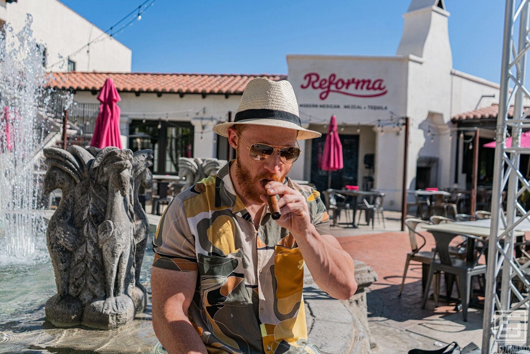 Julian Cisterna smokes a cigar beside a spraying stone fountain, wearing a straw hat, sunglasses, a patterned short-sleeve shirt, and light shorts.