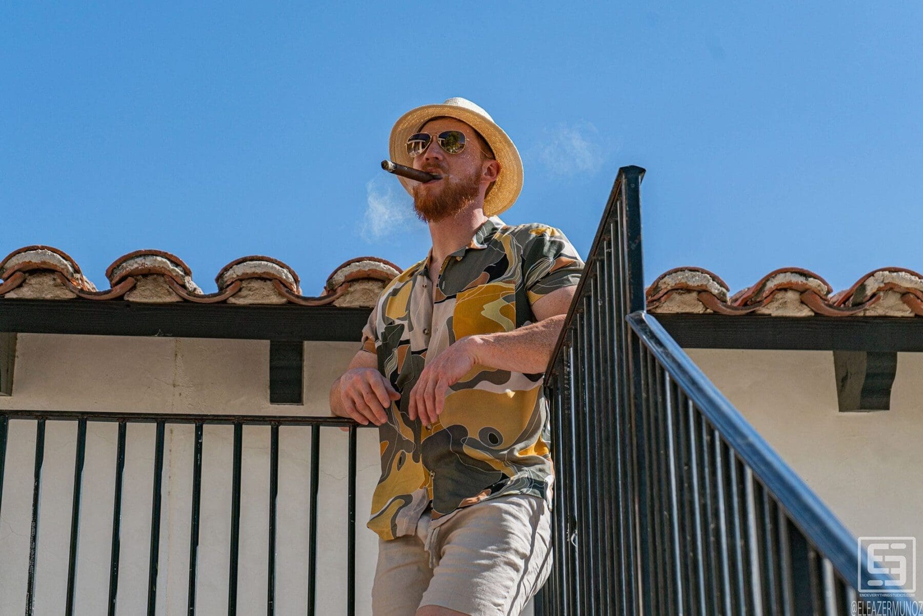 Julian Cisterna stands against a blue sky beside a black stair railing, wearing a straw hat, sunglasses, a patterned short-sleeve shirt, and light shorts.