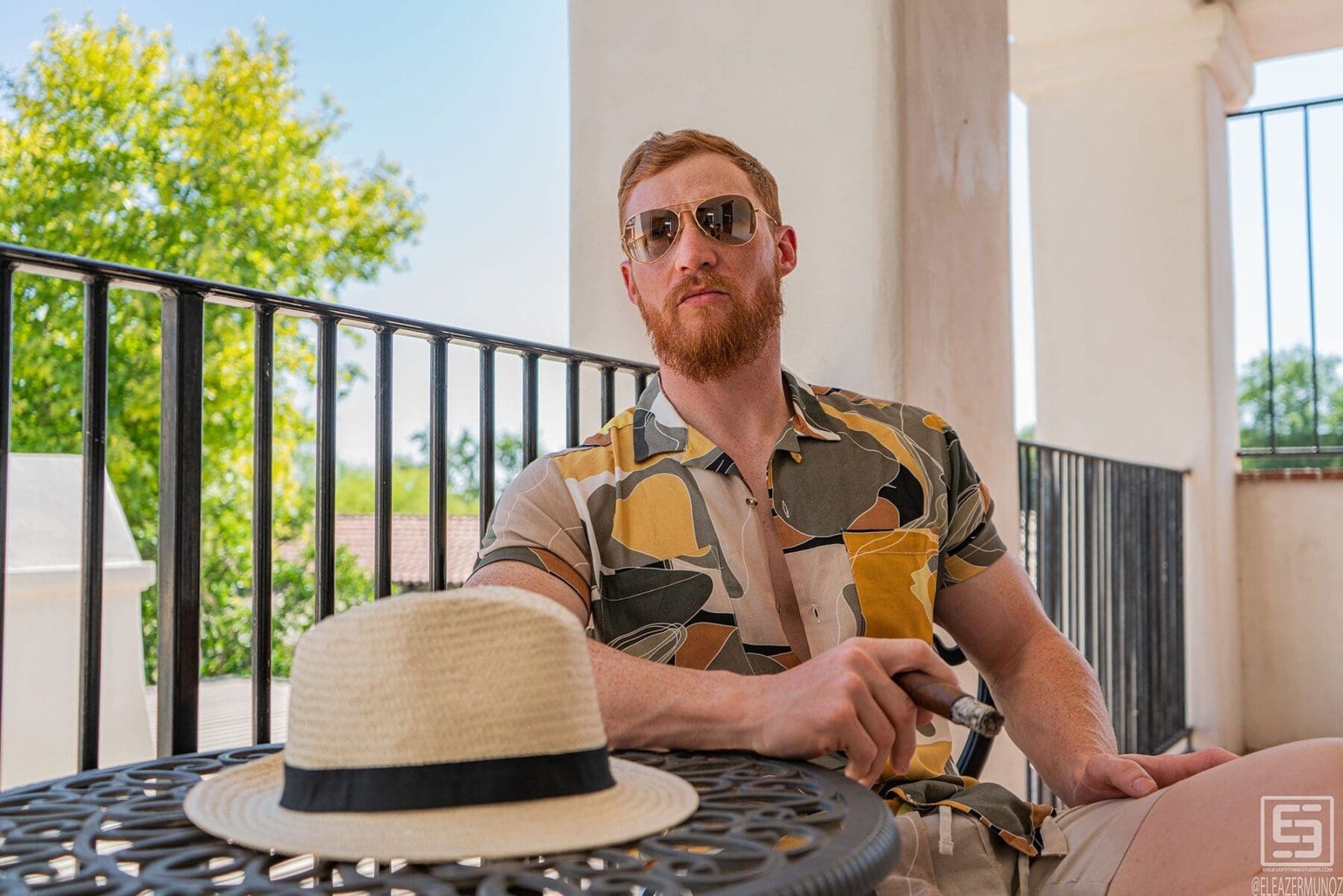 Julian Cisterna sits without the hat and faces the camera beside a round metal table, wearing sunglasses, a patterned short-sleeve shirt, and light shorts.