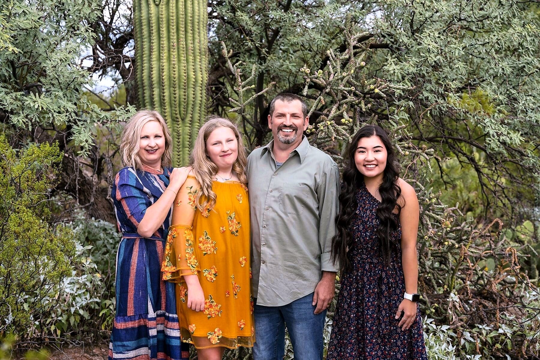 Four adults stand closely together beneath desert trees beside a tall saguaro cactus.