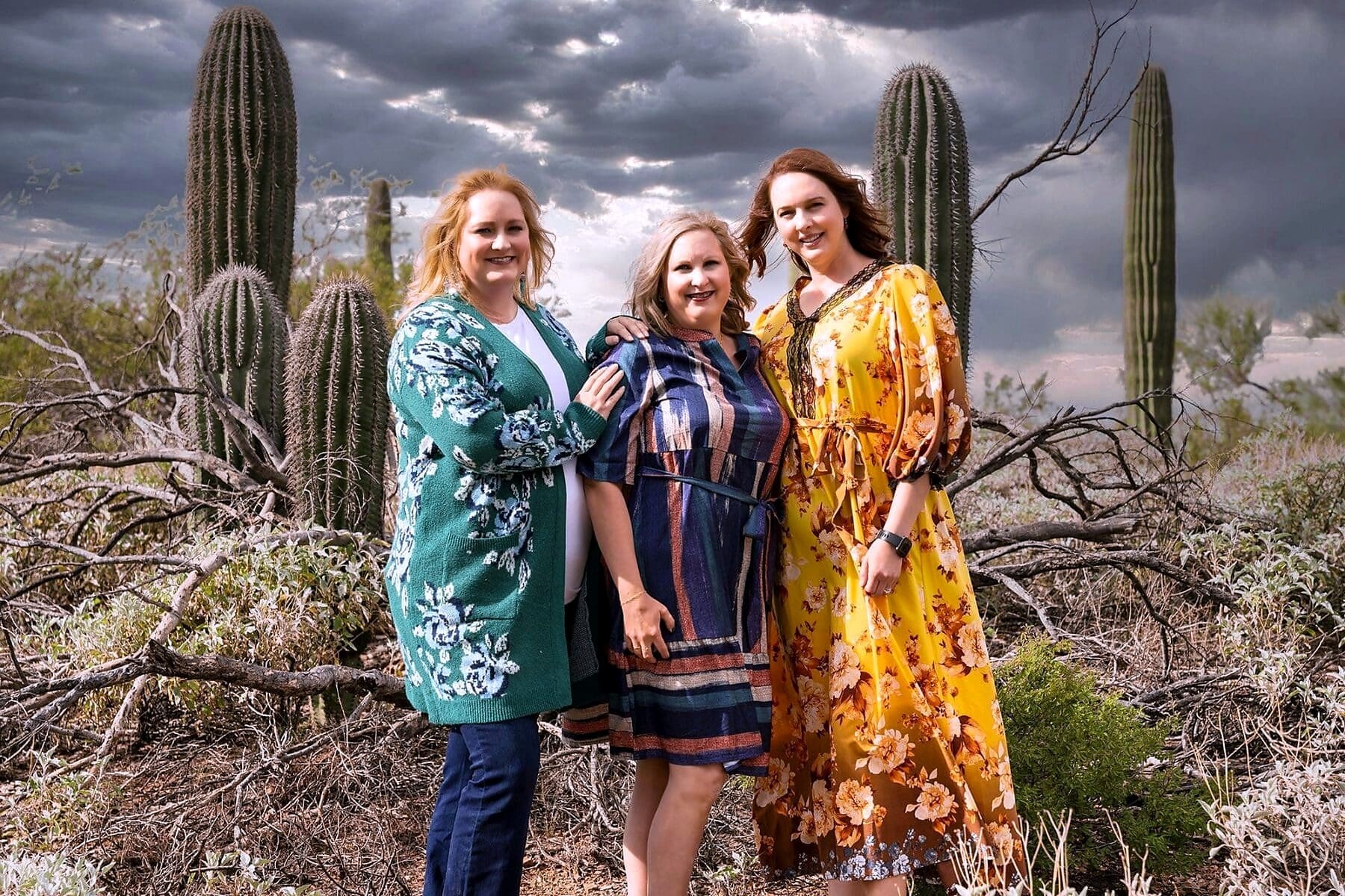 Three women stand closely together among tall saguaros beneath a dramatic cloudy sky.