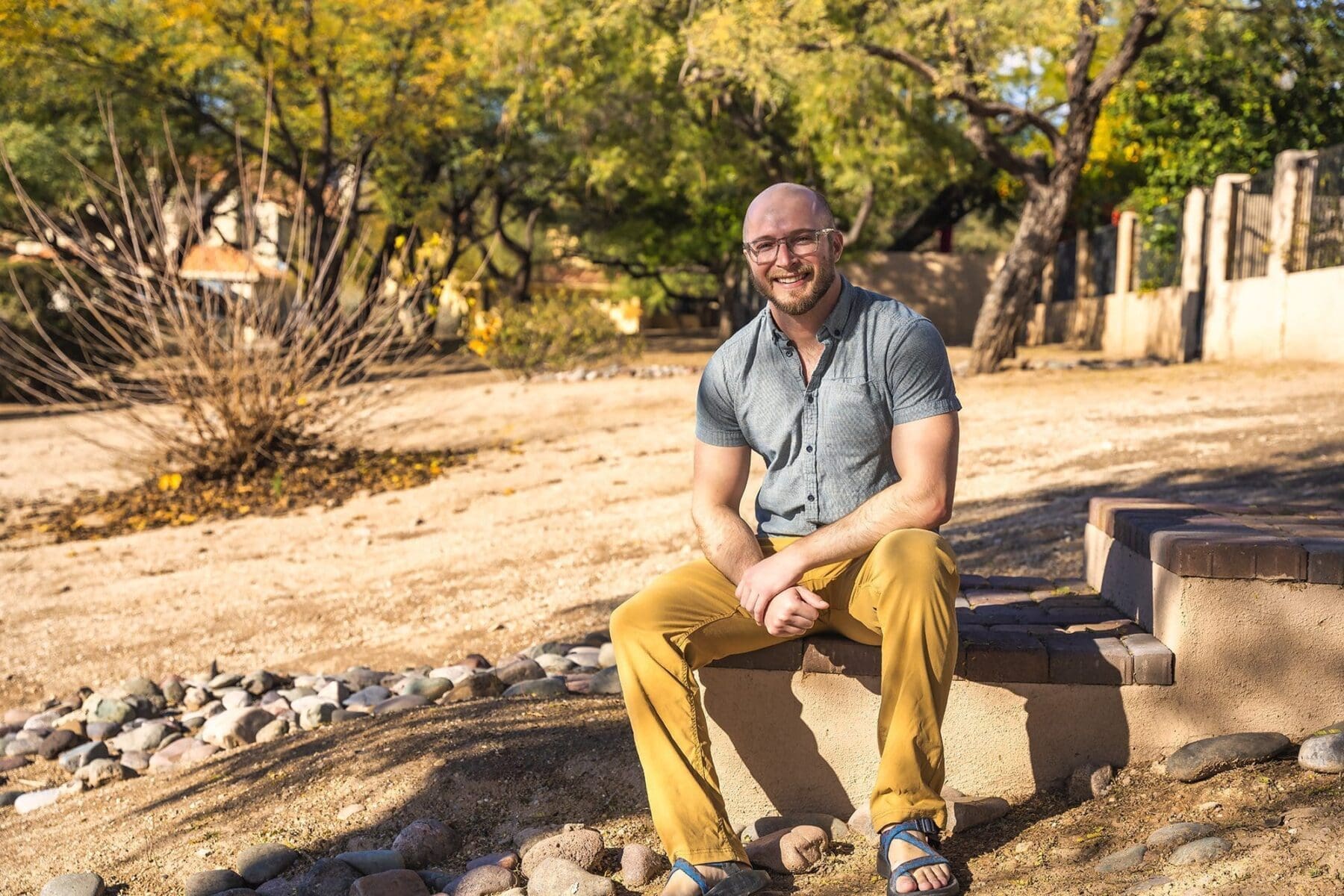 Sean Murphy smiles while seated on a low outdoor ledge in a blue short-sleeve shirt and mustard-colored pants.