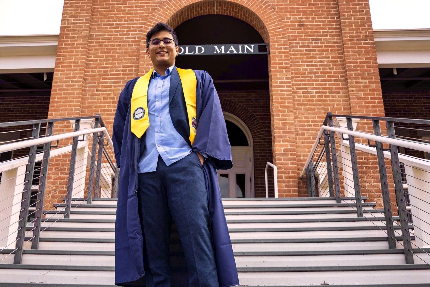 Rehit stands in cap and gown on the stairs below the Old Main sign.
